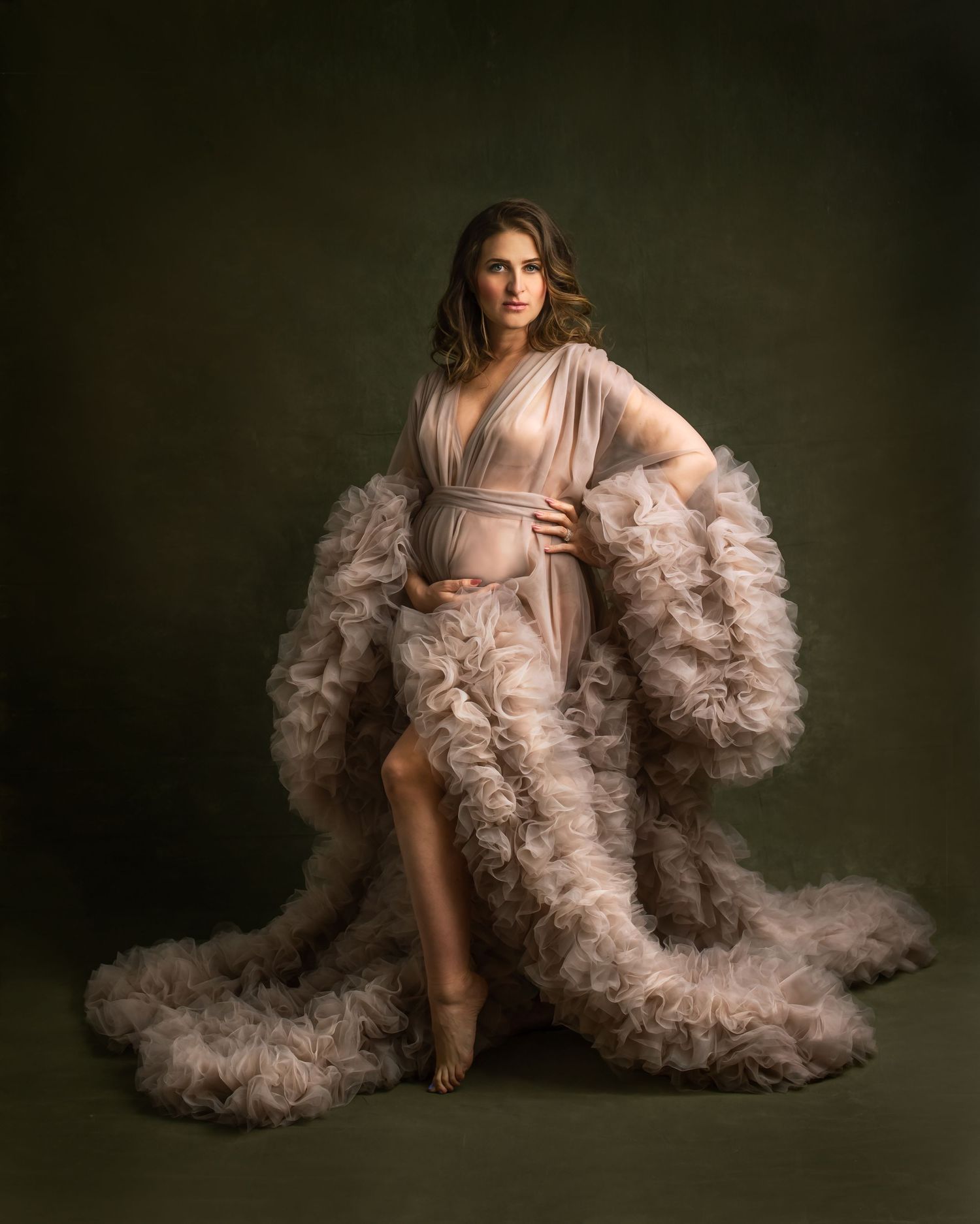 Pregnant woman in elegant ruffled tulle maternity gown posing against dark green studio backdrop.