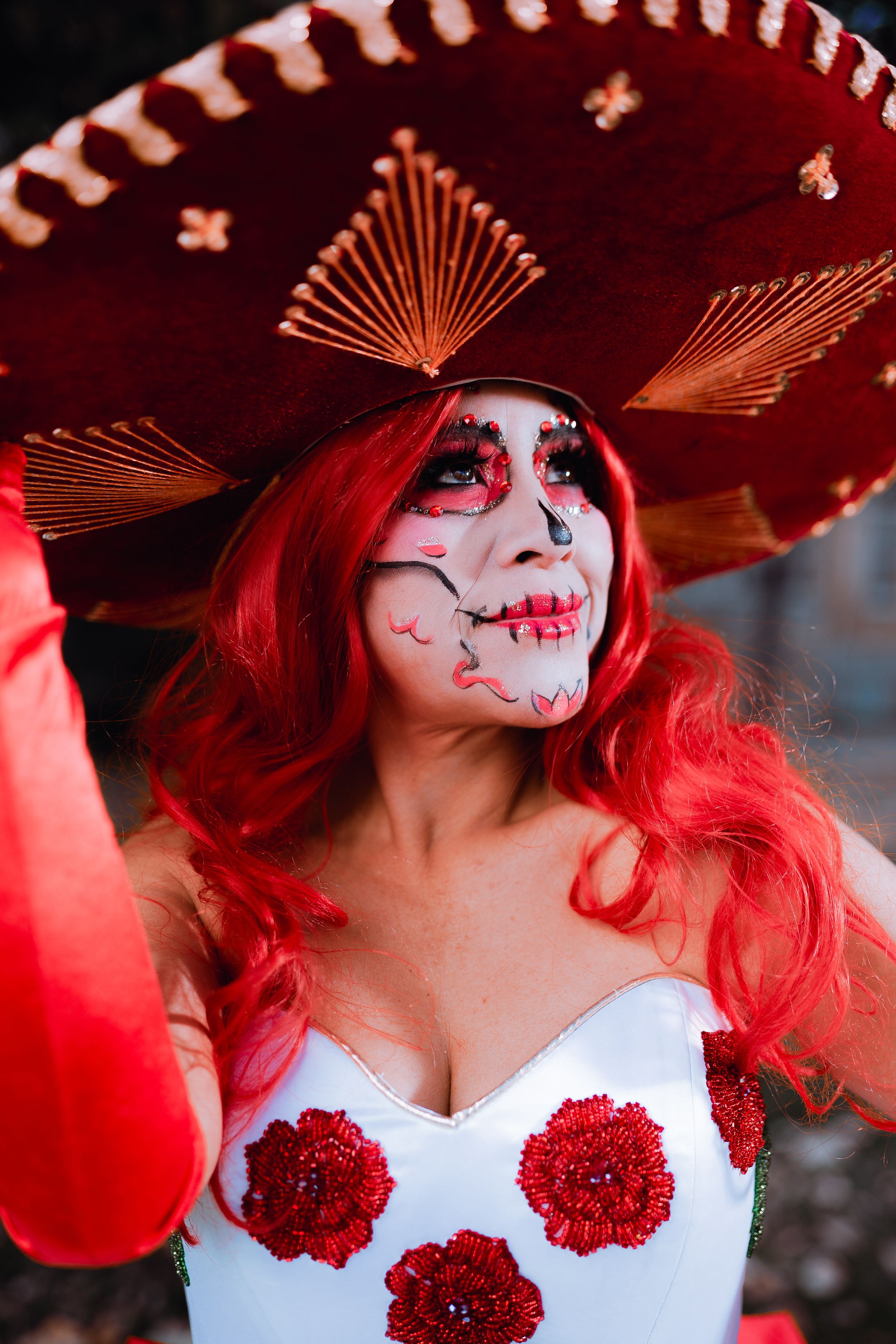 Close-up of Day of the Dead makeup under red sombrero with white dress adorned with red flowers.