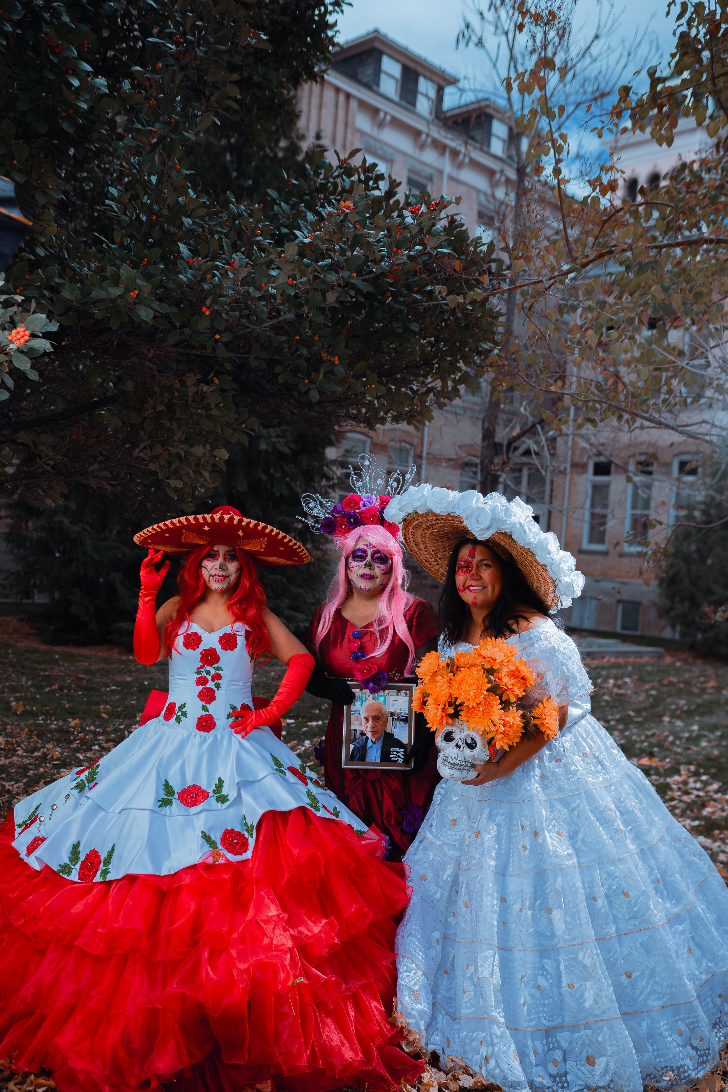 Three people in elaborate Dia de los Muertos costumes featuring red and white dresses with decorative hats.