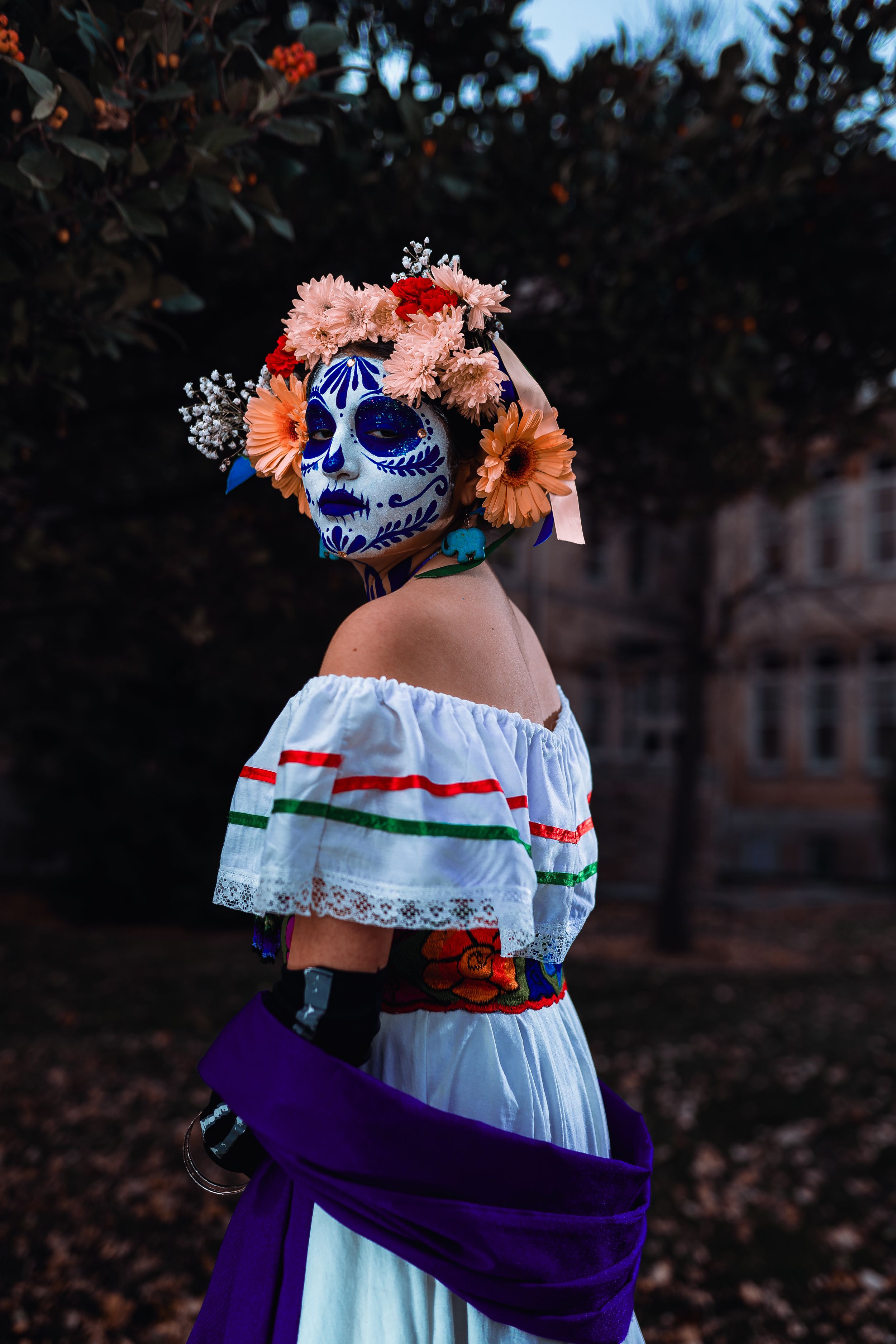 Festive Day of the Dead costume with elaborately decorated sugar skull mask and floral headdress against dark background.