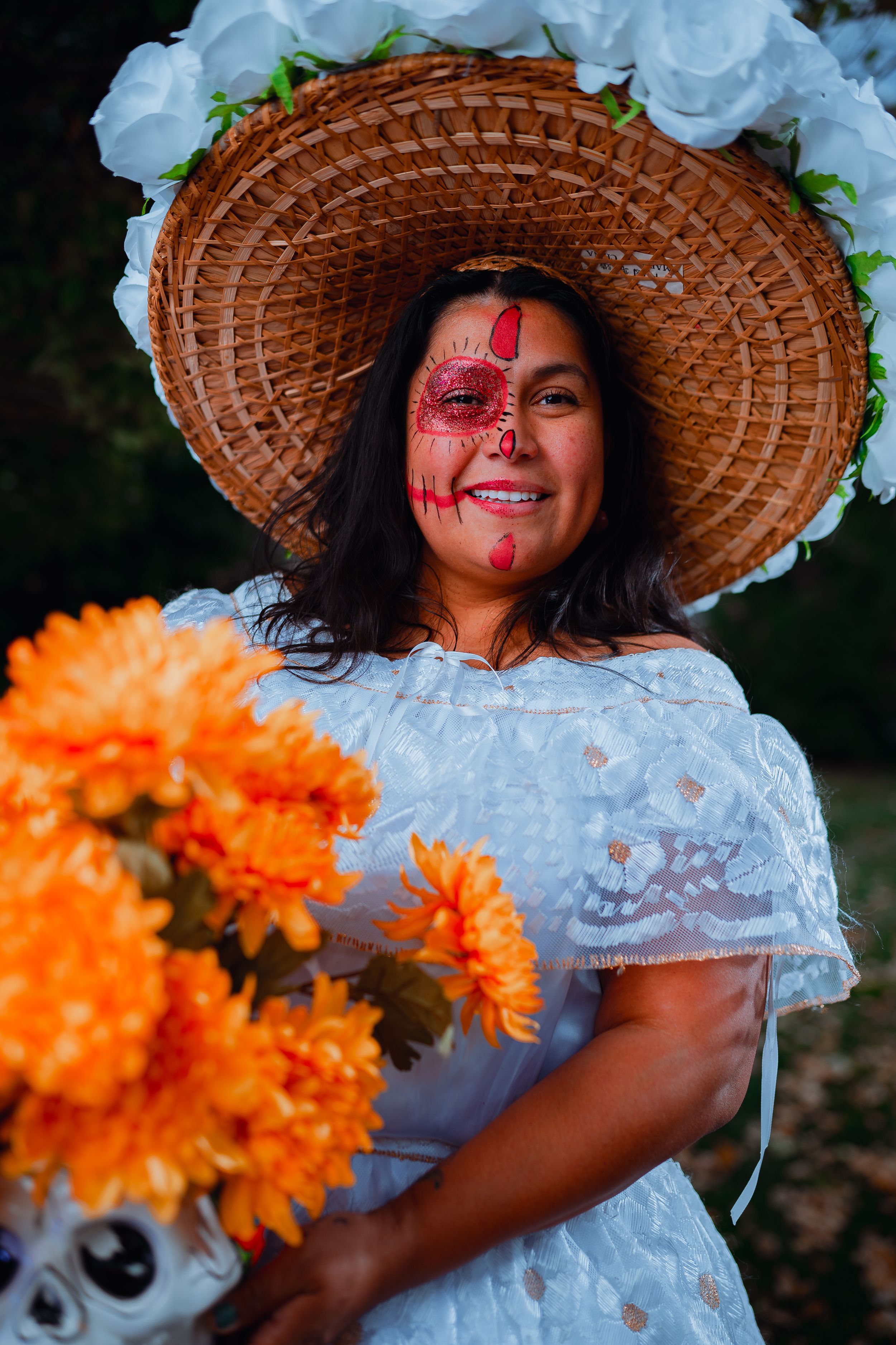 Person in white dress and straw hat with Dia de los Muertos makeup holding orange marigolds and skull vase.