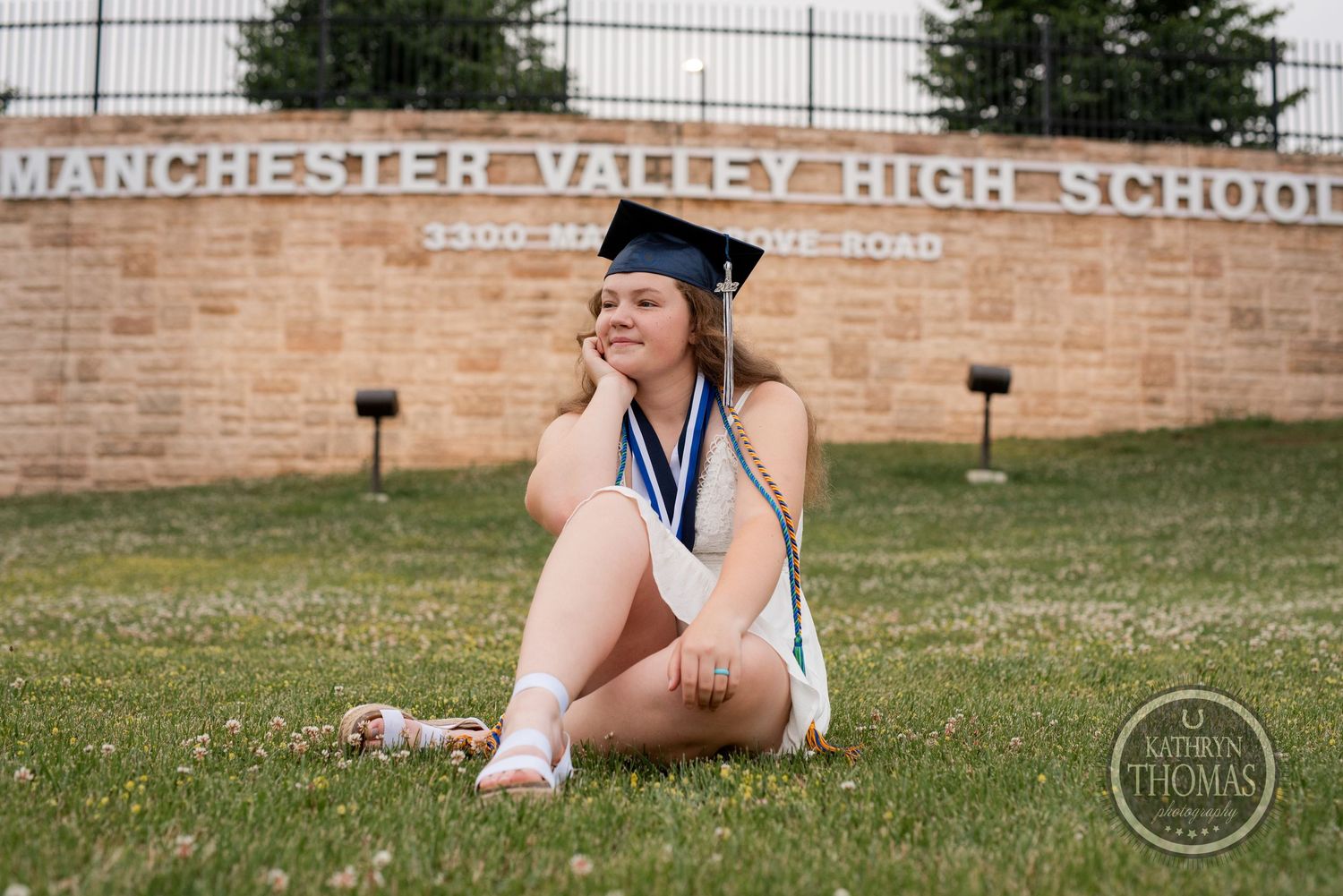 Graduate sits casually on grass in front of school sign during graduation photo session.