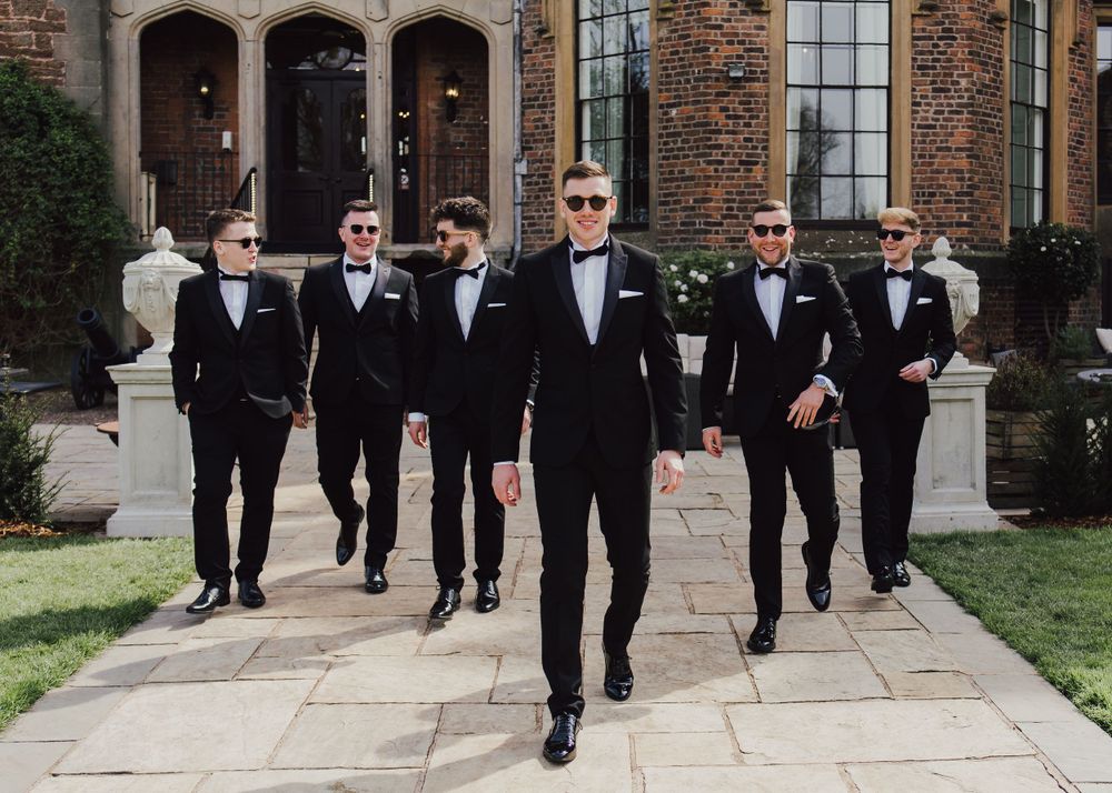 Groomsmen in black tuxedos walk confidently down stone path at elegant brick venue.