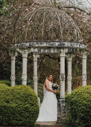 Bride in white gown standing beneath ornate stone gazebo in a manicured garden.