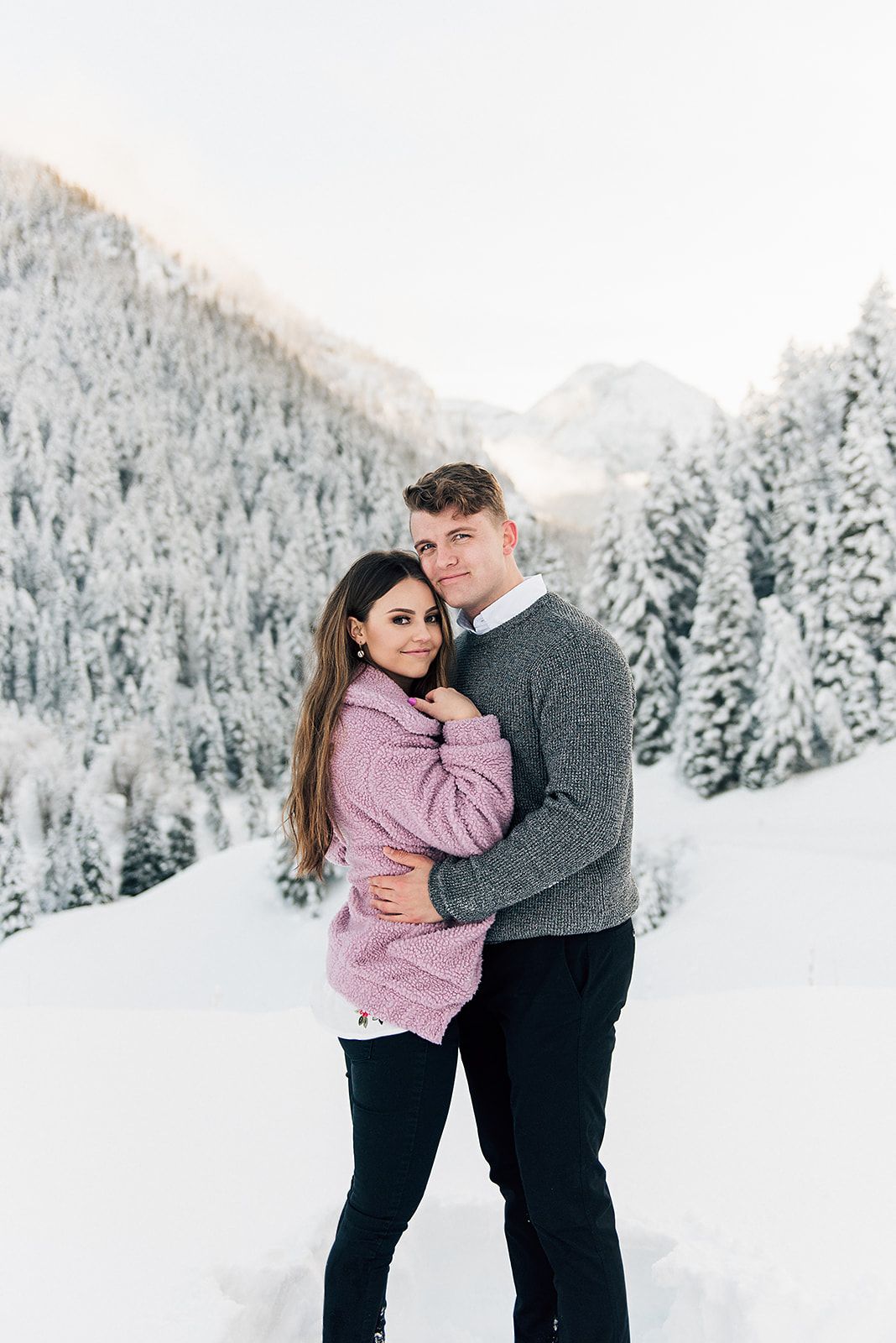 Winter engagement photo with couple in cozy sweaters against snowy mountain vista.