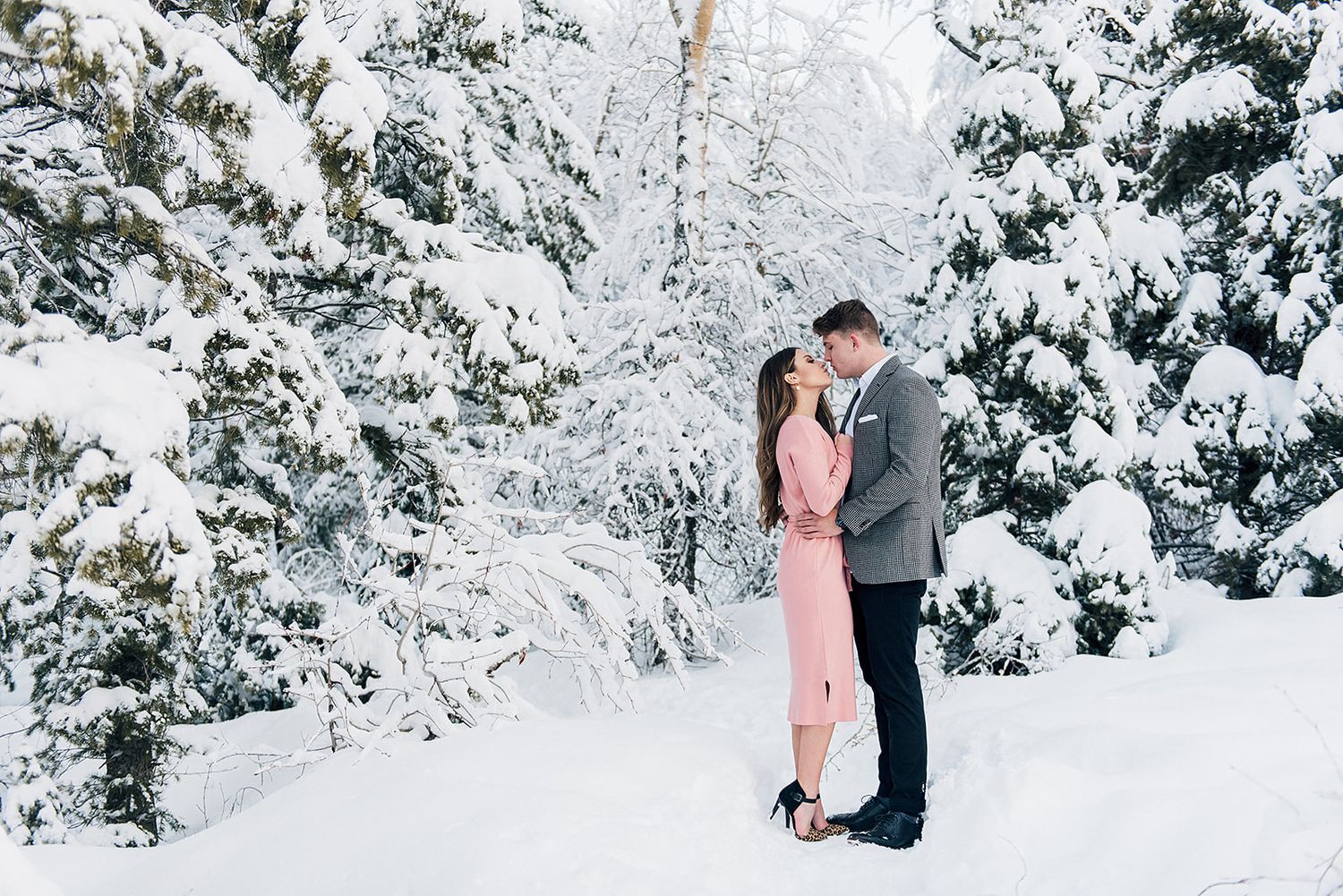 Romantic winter engagement photo with snowy pine trees in background.