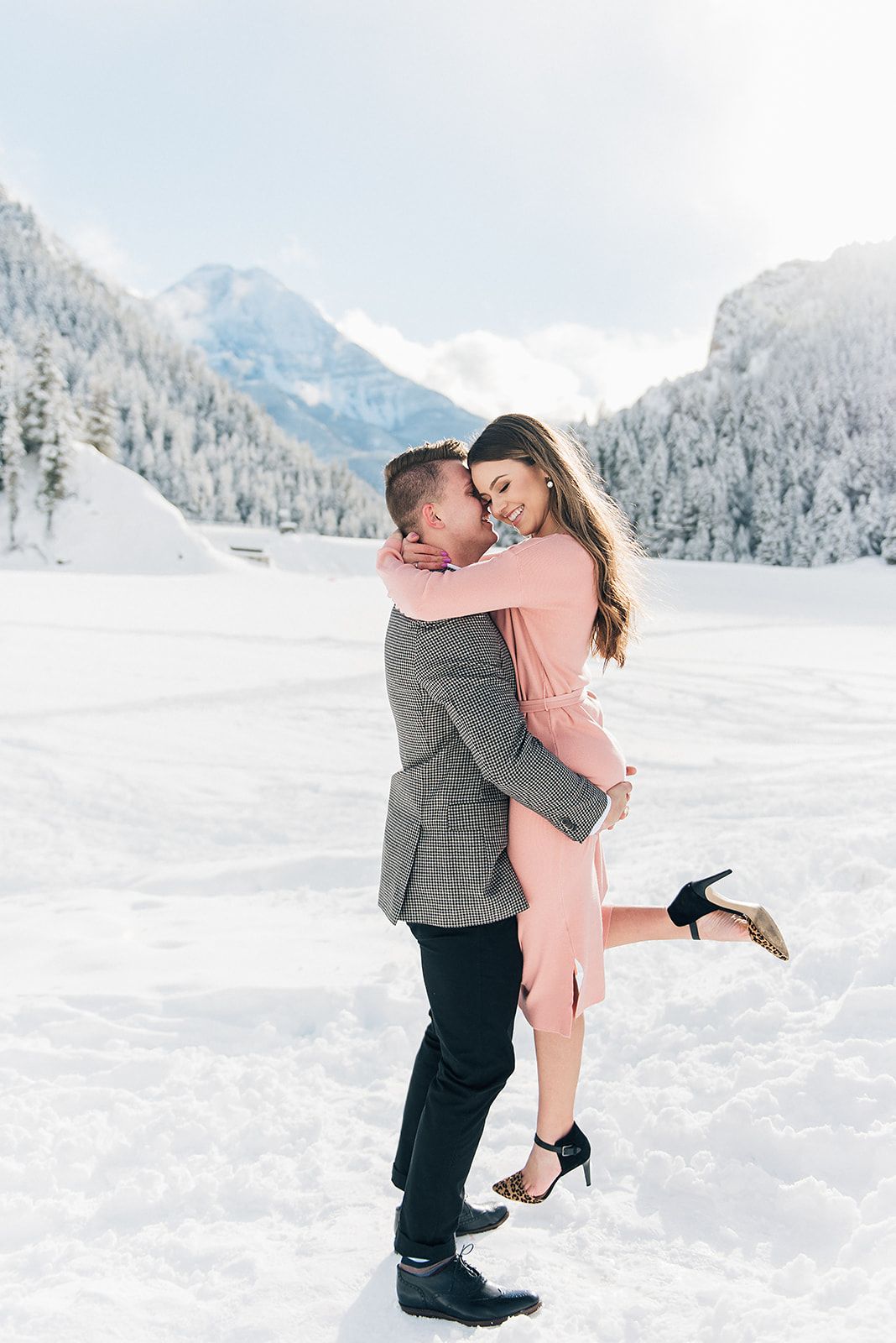 Playful winter engagement photo with snow-capped mountains and pine forest backdrop.