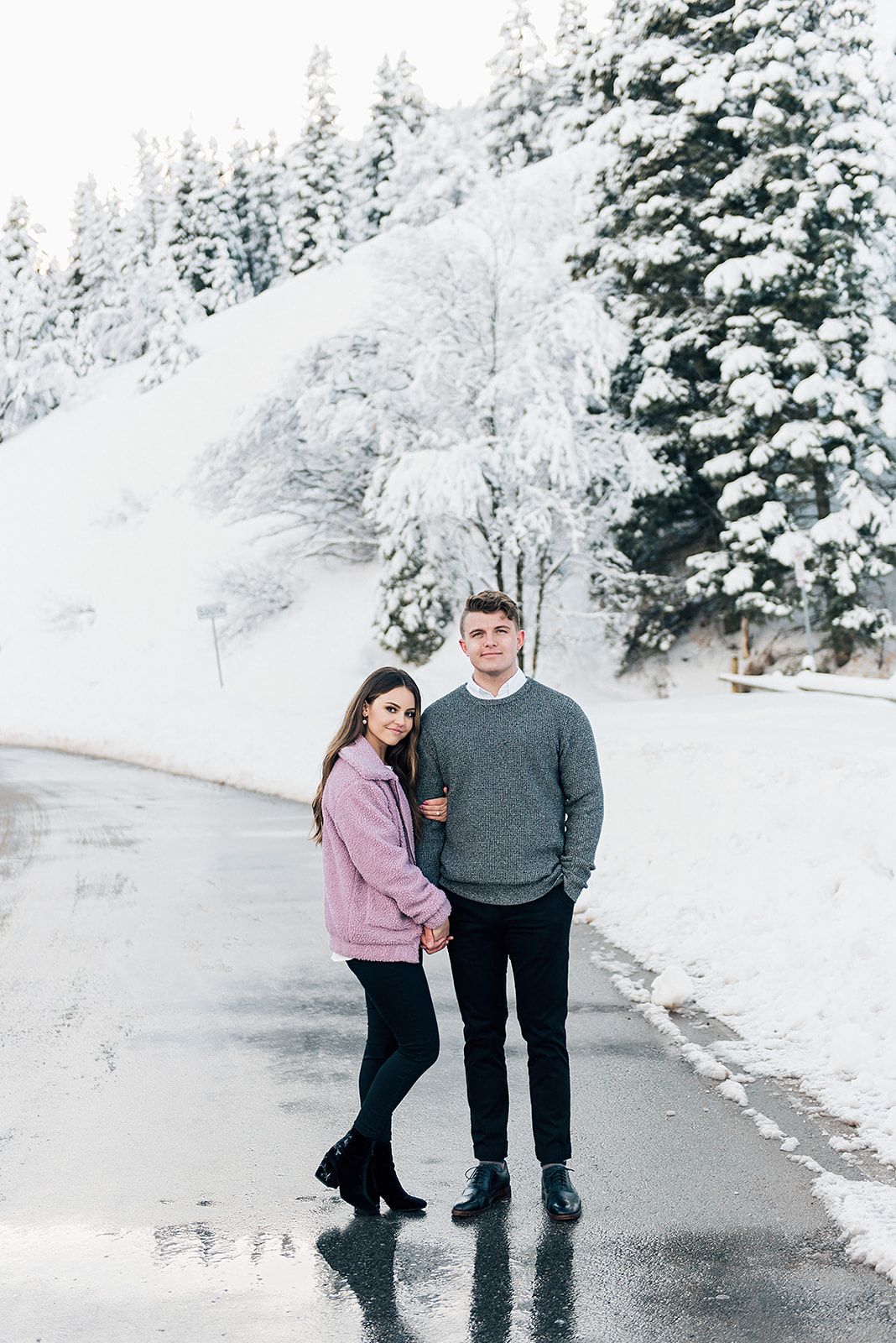 Couple in winter wear stands together on snowy mountain road surrounded by snow-covered evergreens.