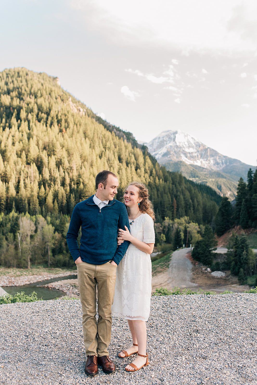 A couple in casual dressy attire poses together with snowy mountain peaks and evergreen forest behind them.