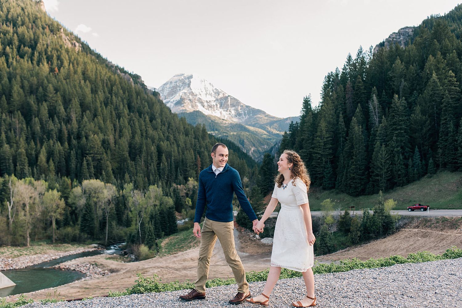 A couple walks hand in hand on a mountain path with dramatic alpine scenery in the background.