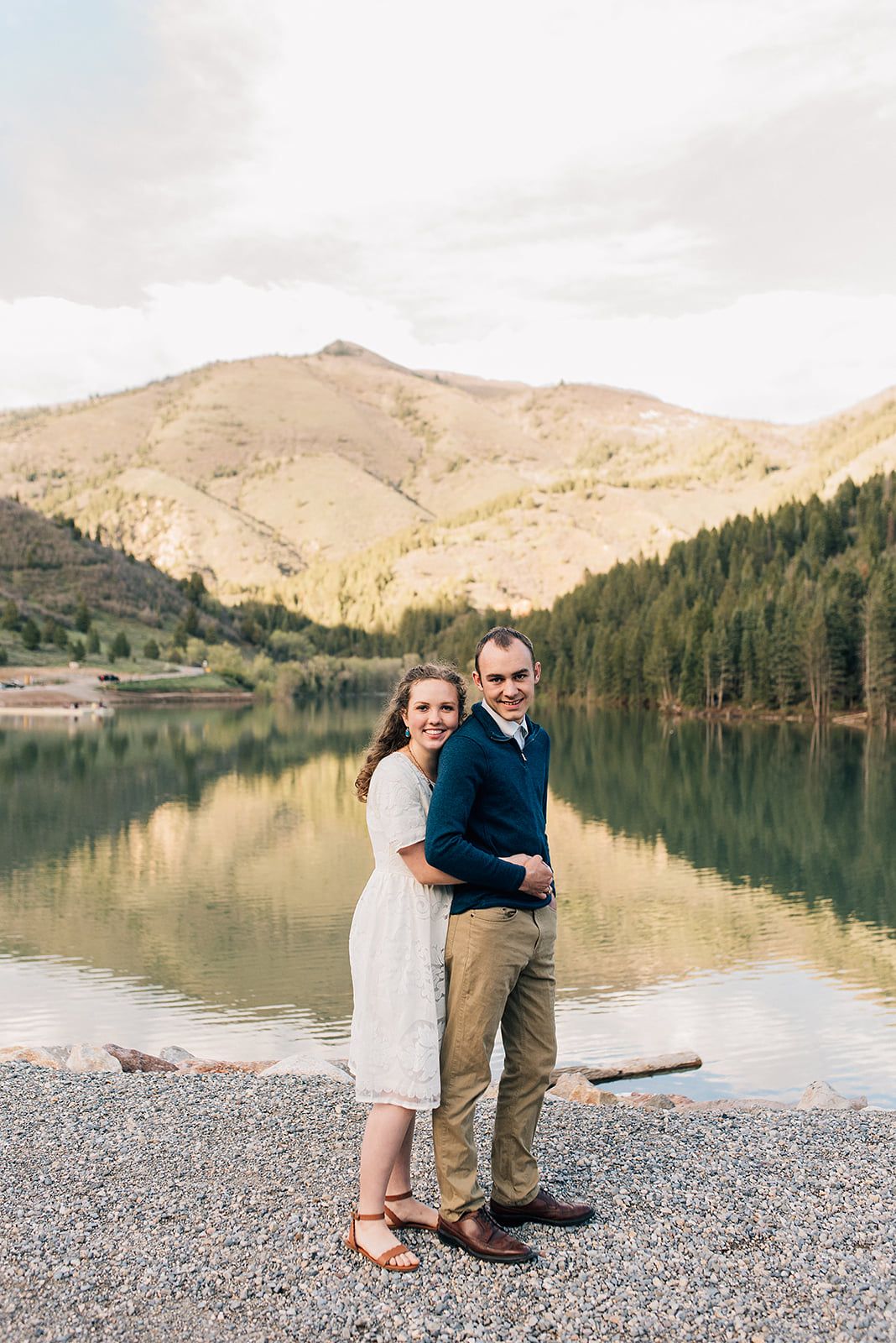 A couple stands together by a serene mountain lake surrounded by forested hills and mountains.