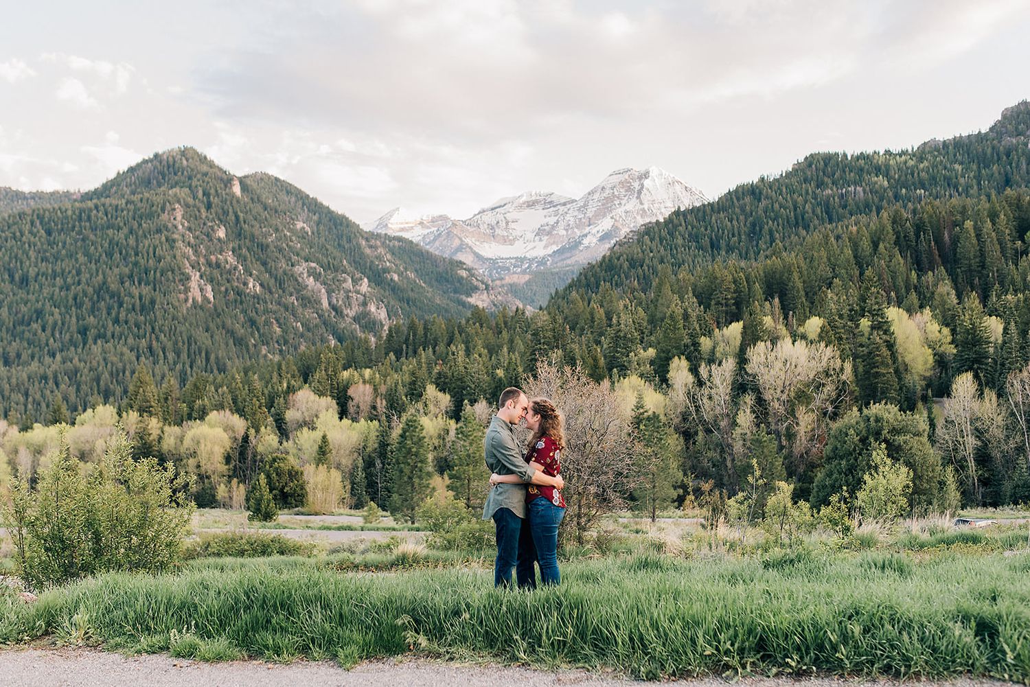 A couple embraces in a mountain valley with dramatic peaks and evergreen forests in the background.