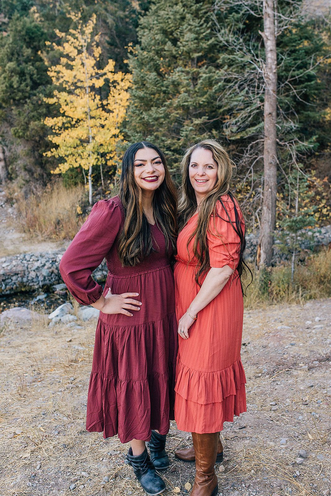 Two friends in flowing burgundy and coral dresses pose against autumn foliage in a mountain setting.