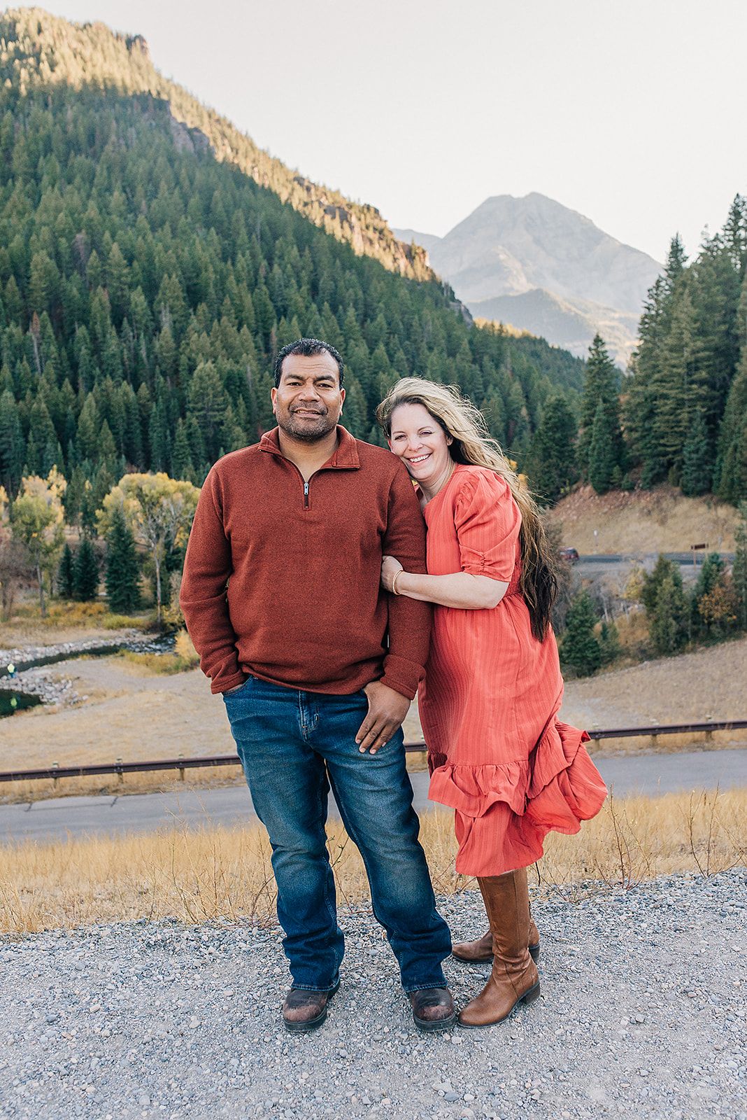 Couple in casual fall attire poses on mountain trail with scenic forest backdrop.