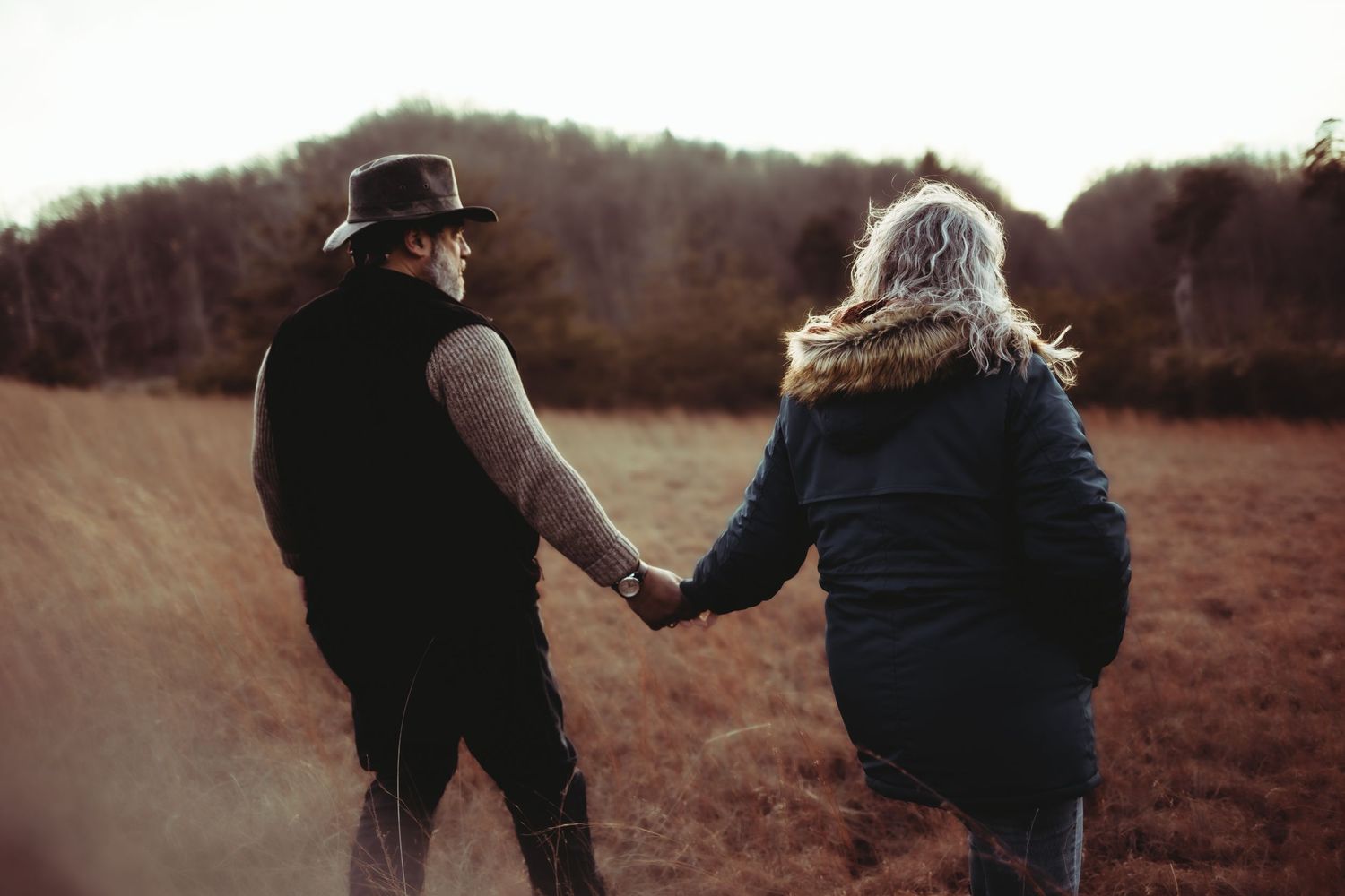 A couple holds hands while walking through an autumn field at sunset.