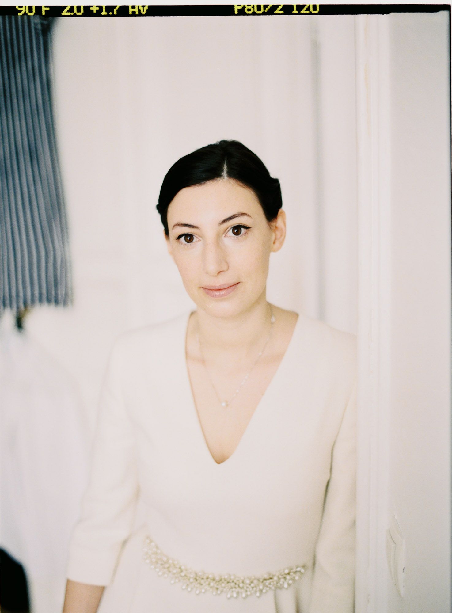 Woman in white v-neck dress with beaded waistline detail against light background.