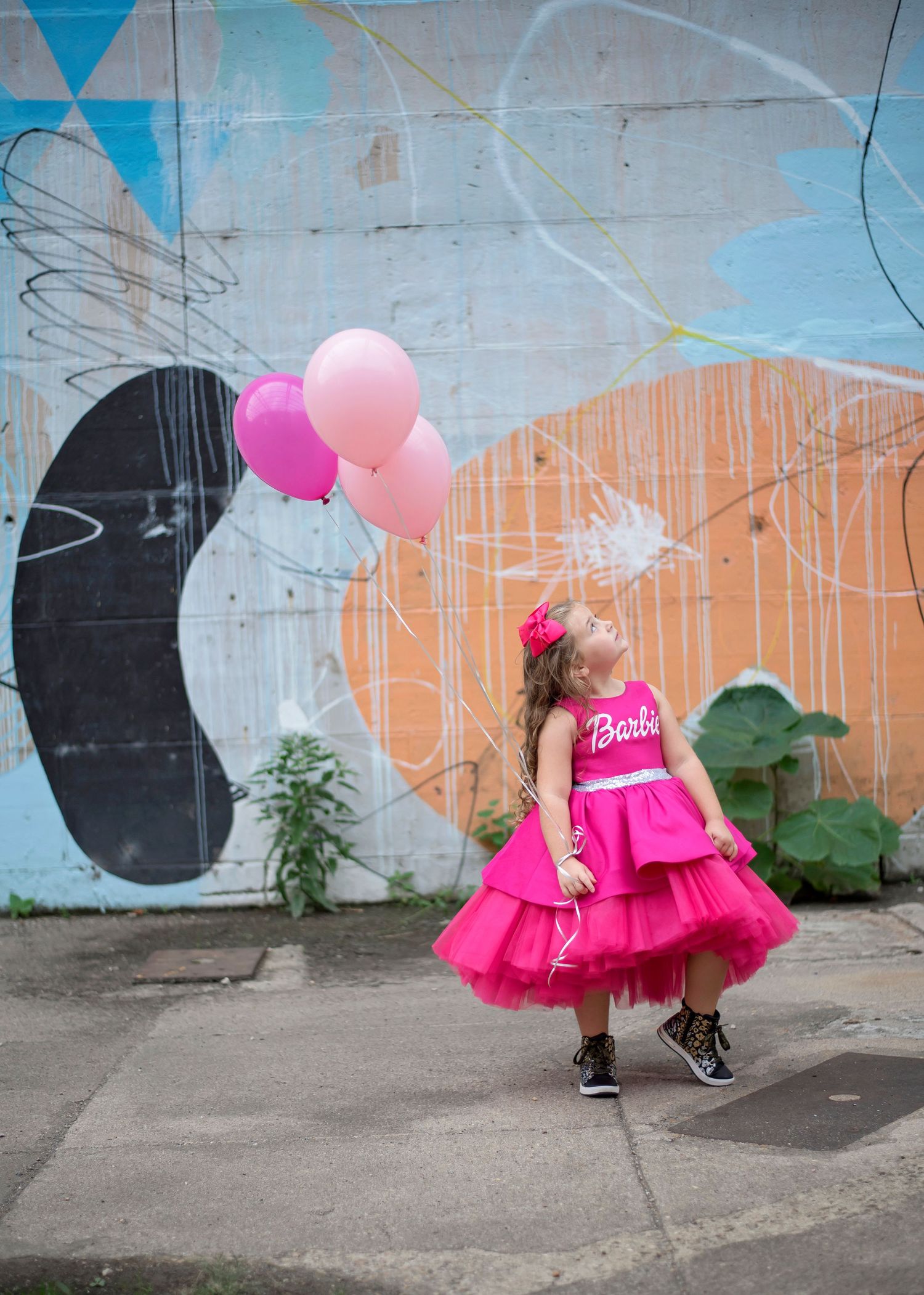 A child in a bright pink tulle dress twirls with balloons against a colorful graffiti wall.