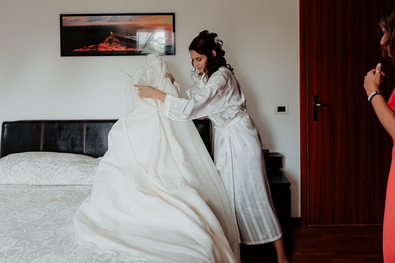 A bride in a white robe preparing her wedding dress on a bed.