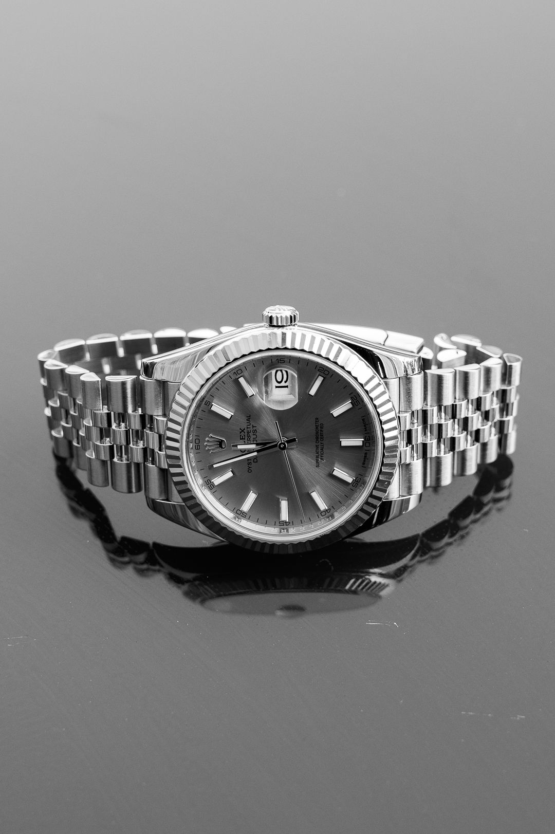 Silver Rolex watch with fluted bezel and jubilee bracelet reflected on black surface.