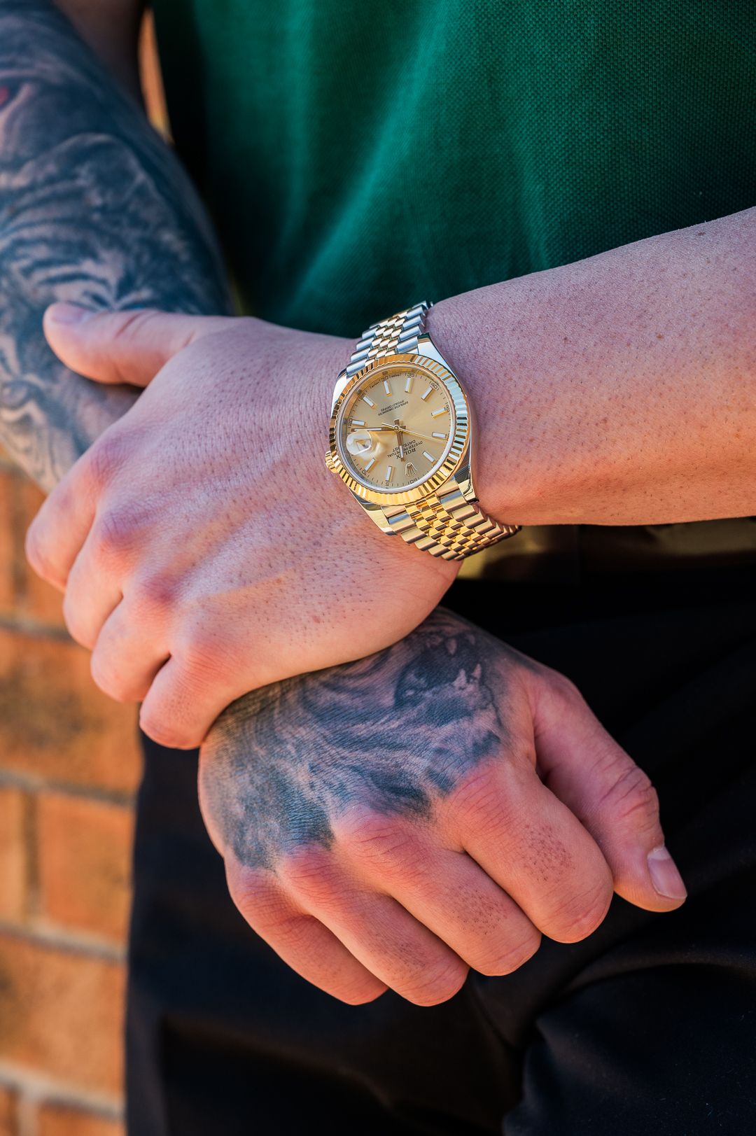 Rolex worn on tattooed wrist against brick wall background.