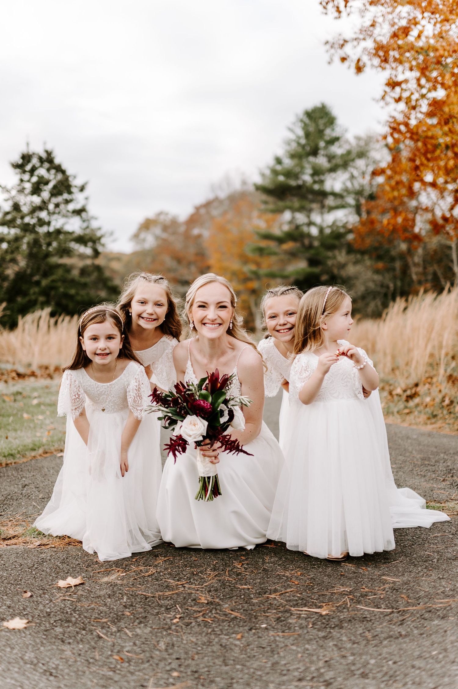Flower girls in white dresses sit together laughing on gravel path with autumn scenery.