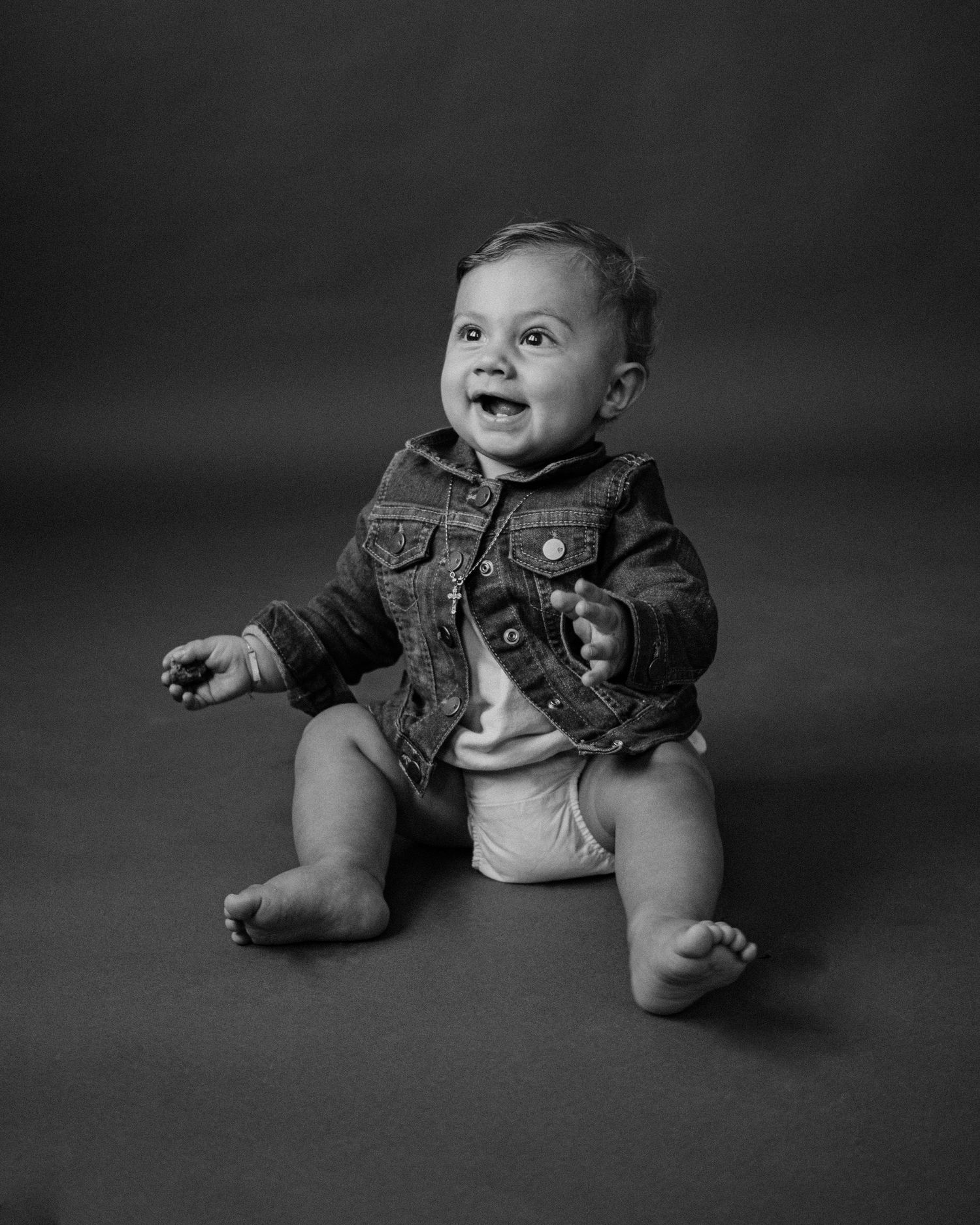 Studio portrait of baby in denim jacket sitting against dark background.