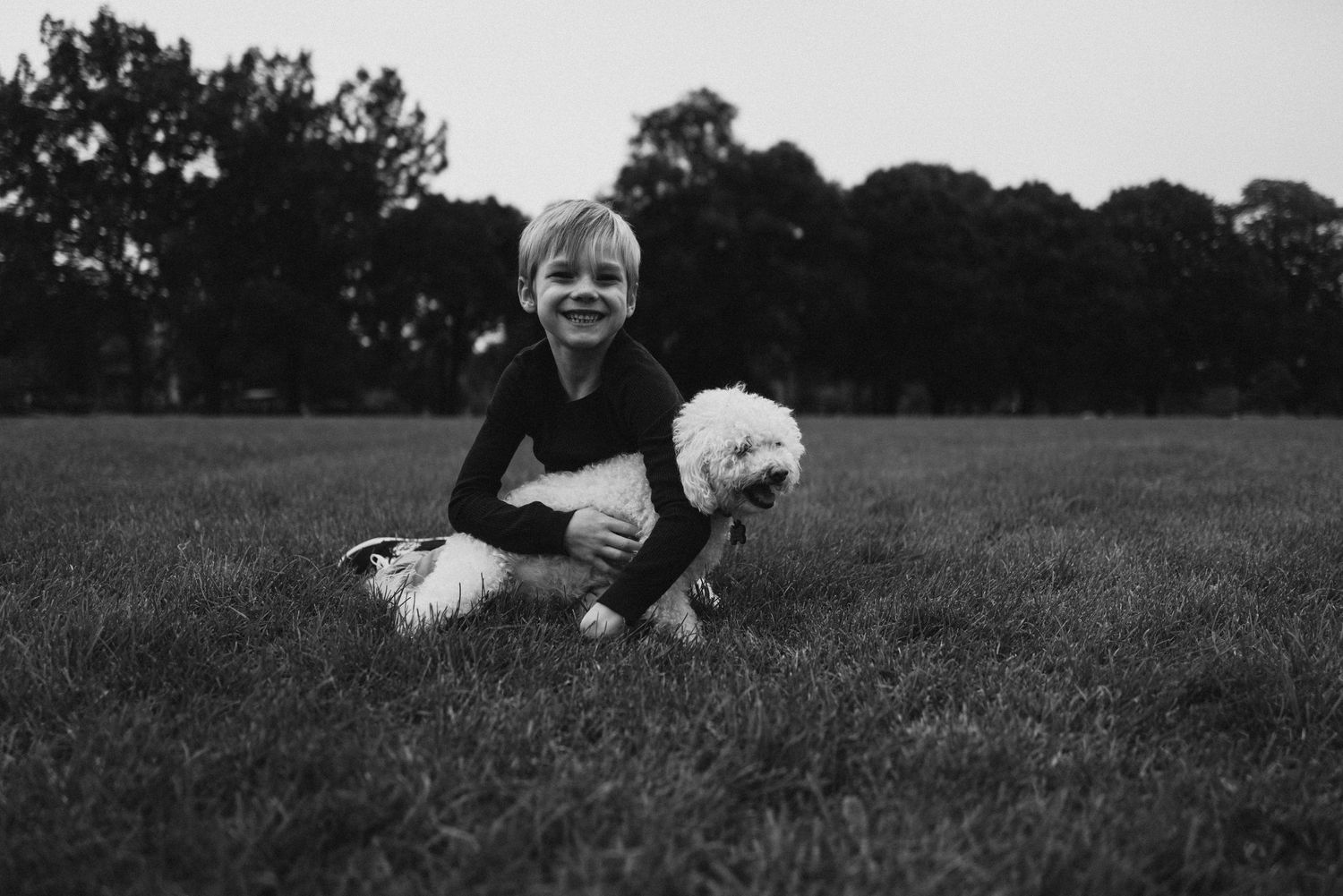 Black and white photo of child sitting on grass with small white dog.