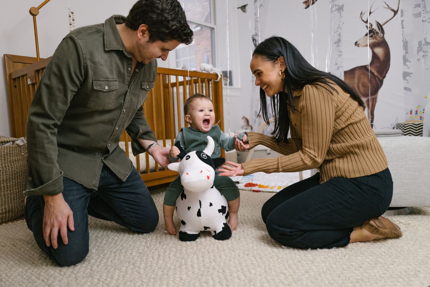 Parents sitting on carpet playing with happy toddler and stuffed animal in nursery with woodland themed wallpaper.