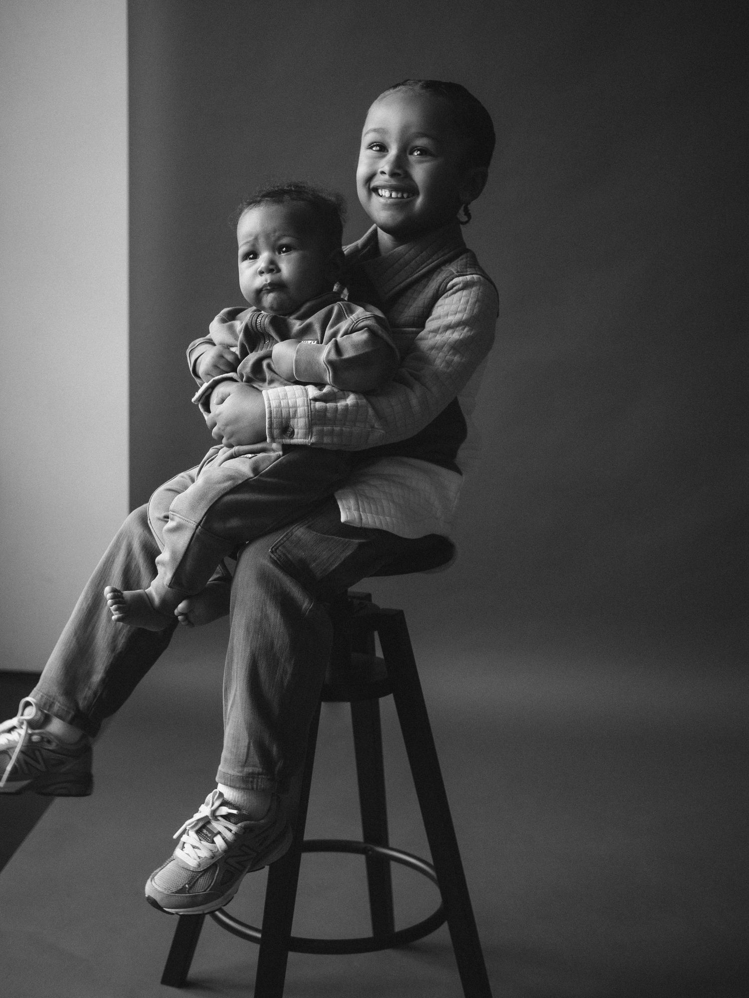 Two children share a tender moment while posing on a bar stool in black and white.