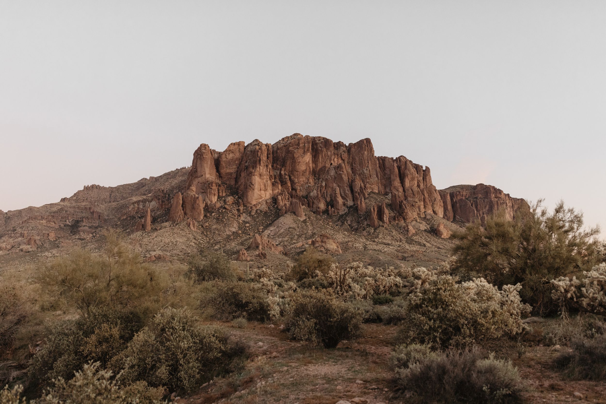 A rugged mountain formation rises above desert terrain in warm golden light. Lost Dutchman mountains, Arizona engagement session location.