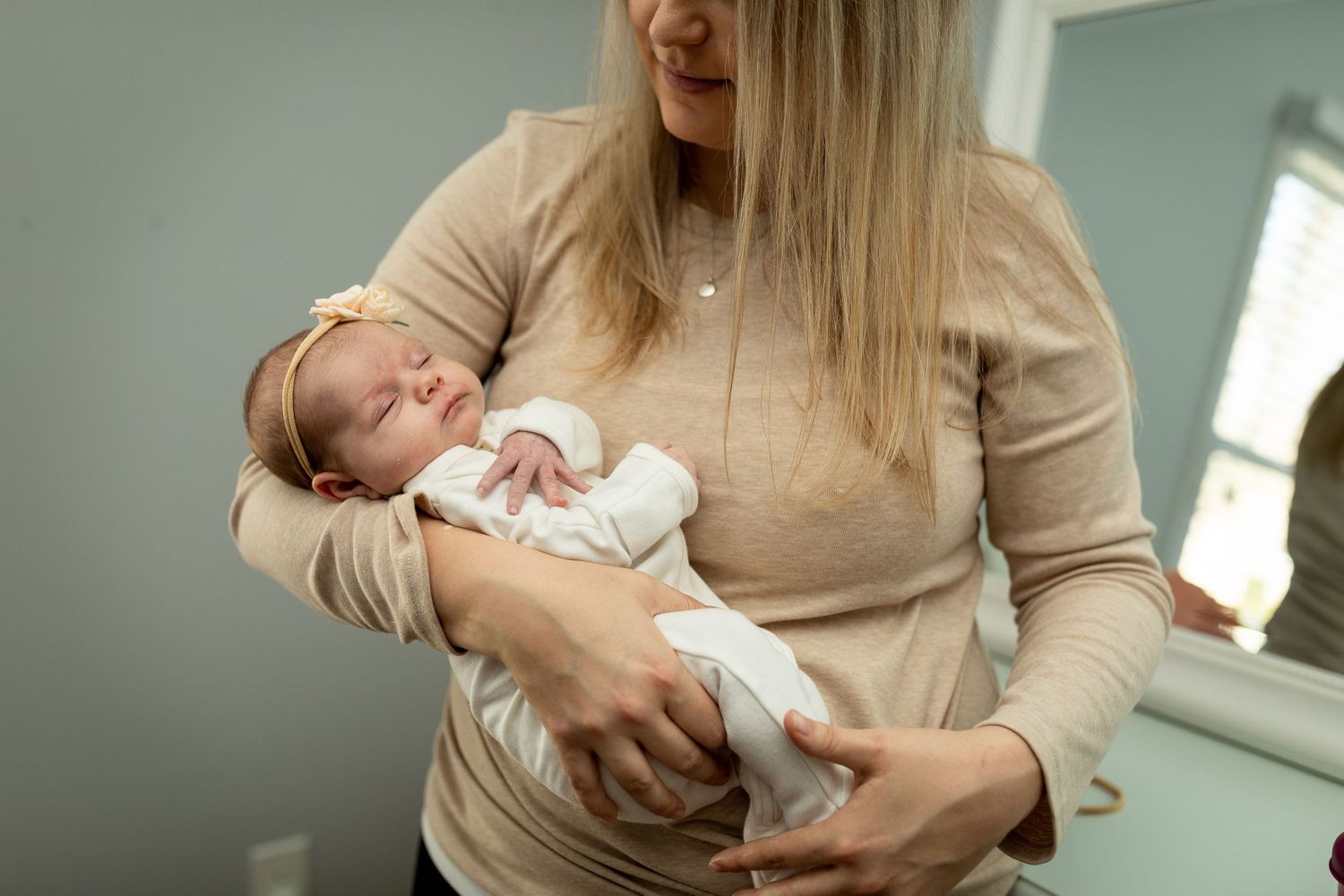 A caregiver in beige sweater gently holds a sleeping newborn in white clothing.