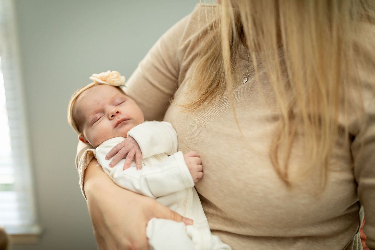Infant in white outfit rests against person wearing beige sweater near window.