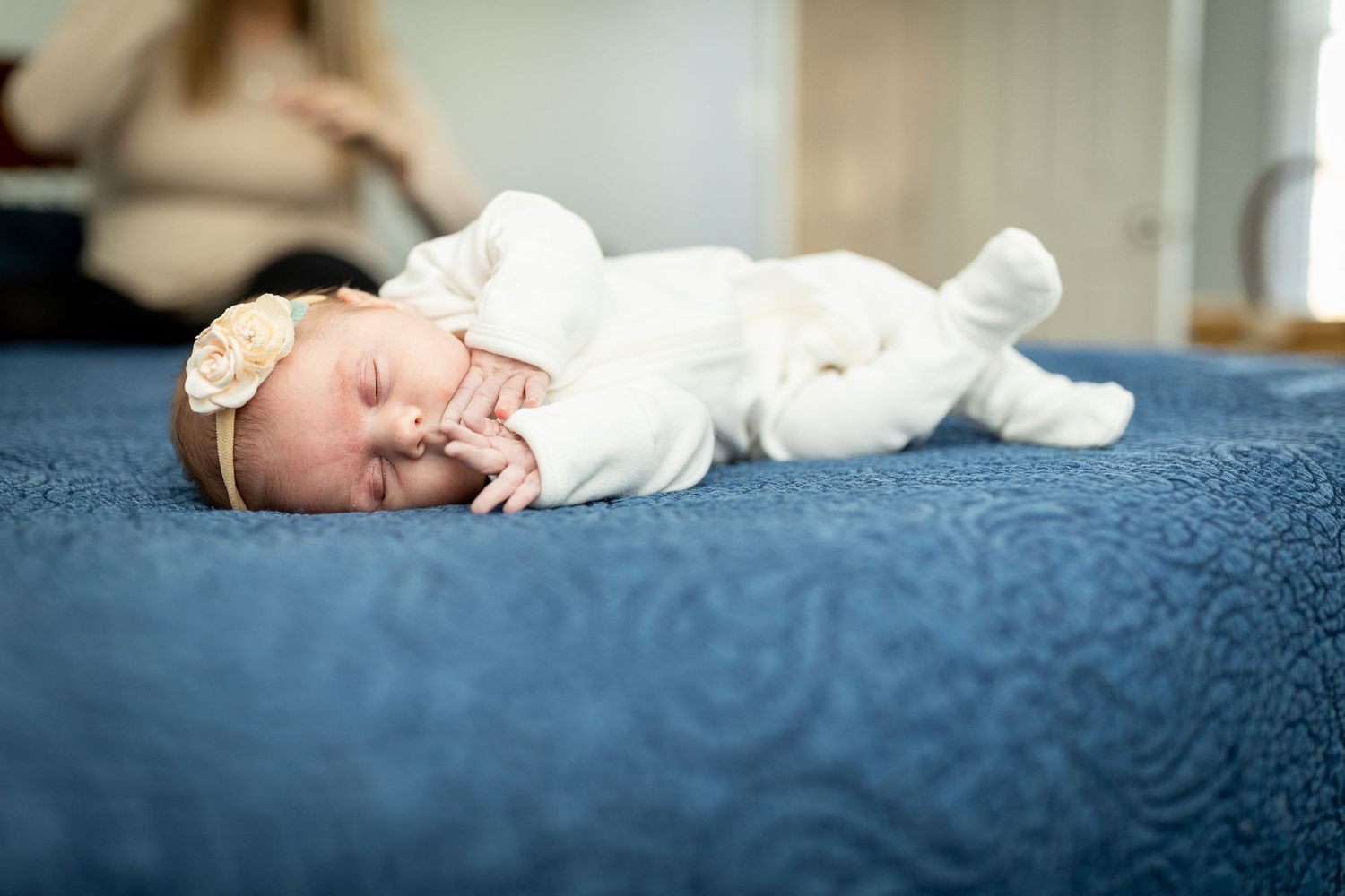Newborn baby wearing white sleeps peacefully on blue bedding while wearing a delicate headband.
