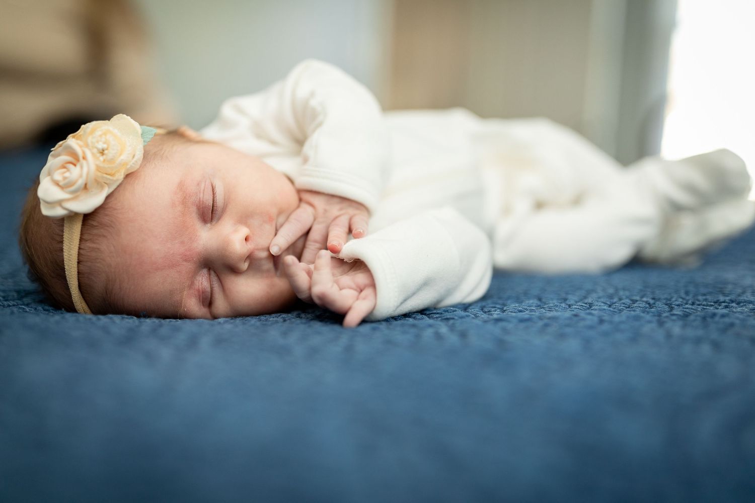 Sleeping infant in white outfit rests on dark blue bedding while wearing beige headband.