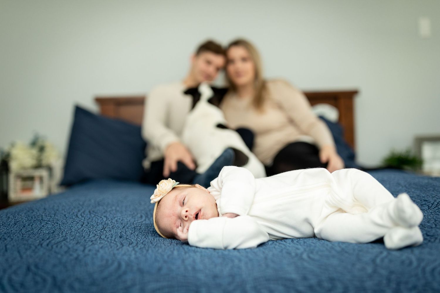 Baby in white sleeps on blue bed while parents sit in background.