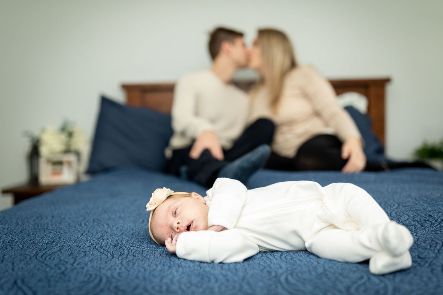 Baby sleeps peacefully in white outfit on blue bedding while parents embrace in background.