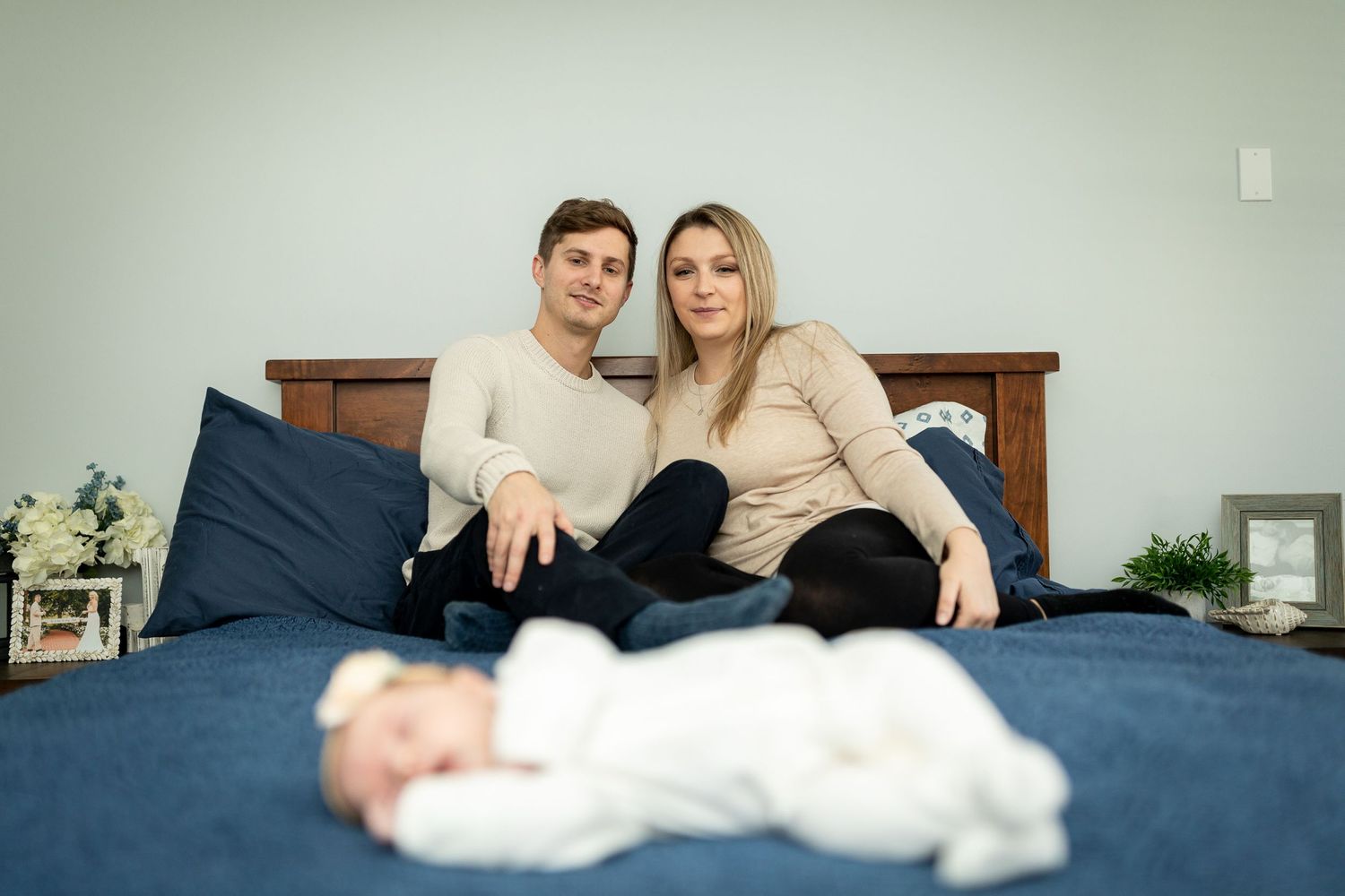 Family portrait on blue bed with baby in white sleepwear in foreground.