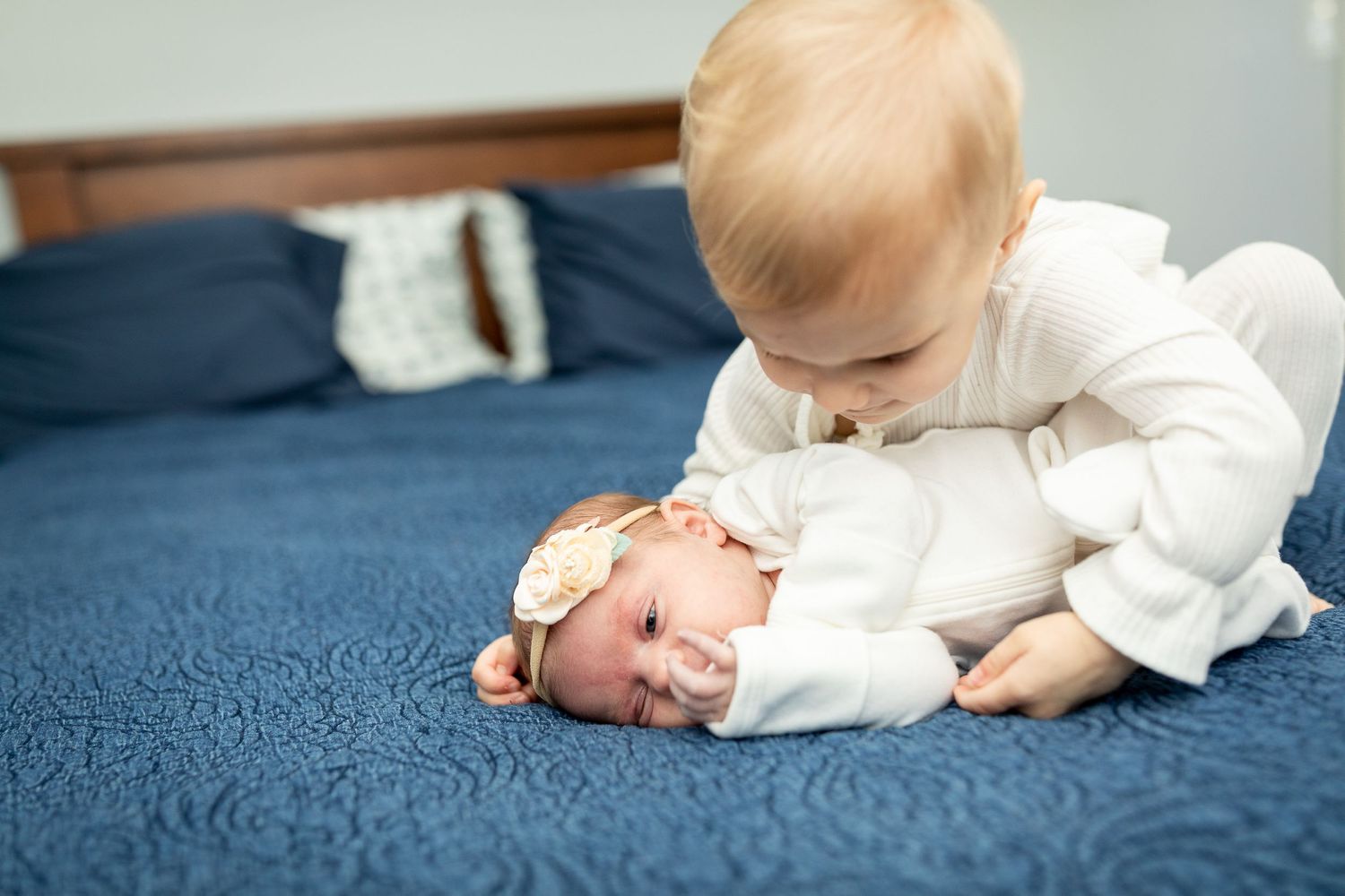 Two infants interact on blue bedding wearing matching white outfits.