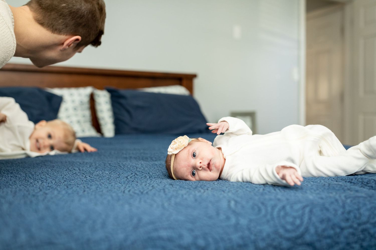 Baby in white outfit rests on blue bedding during family photo session.