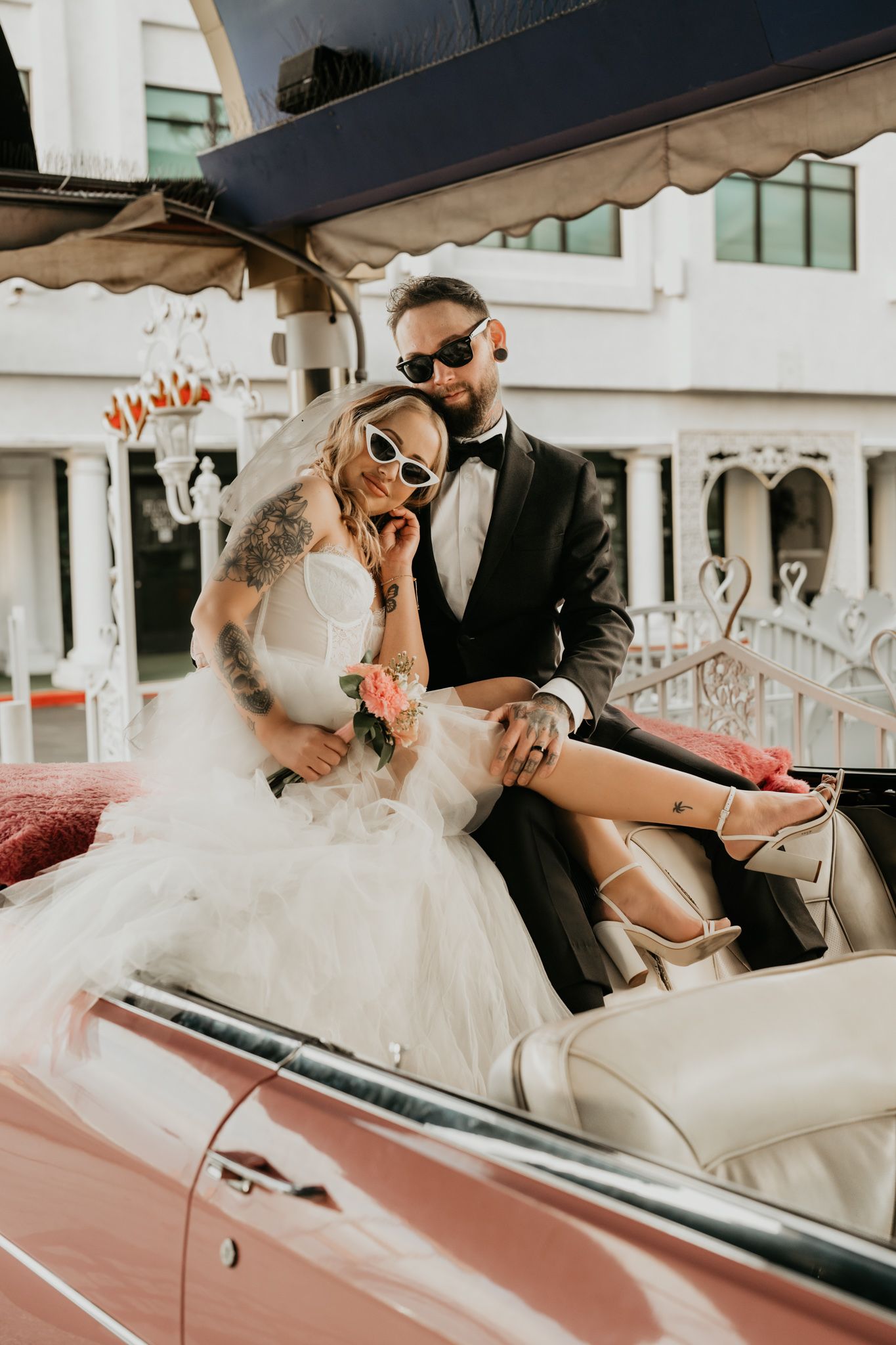 Stylish bride and groom wearing sunglasses pose in classic convertible car.