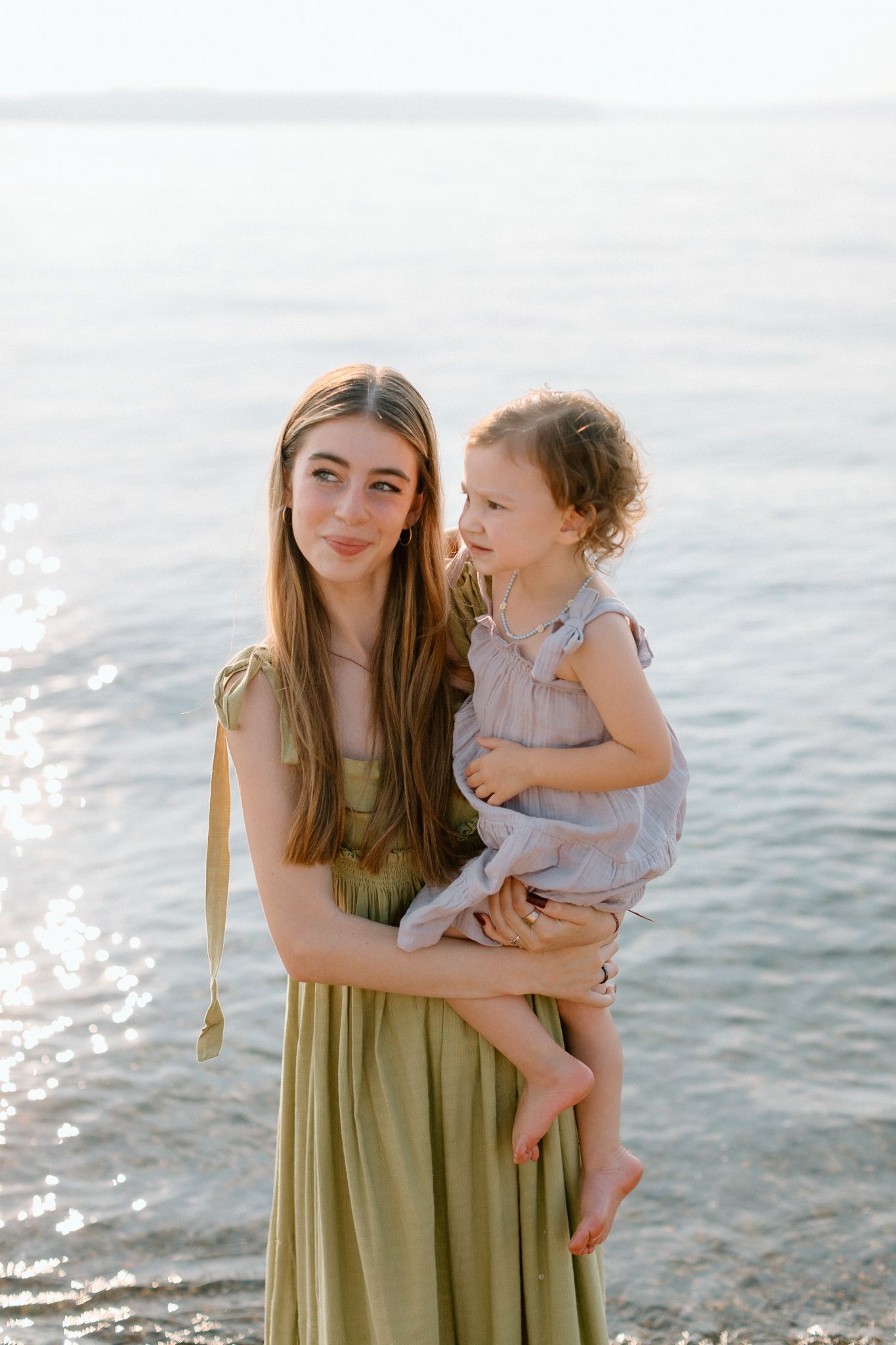A young aunt in an olive green dress holds her toddler daughter by the sparkling sea.