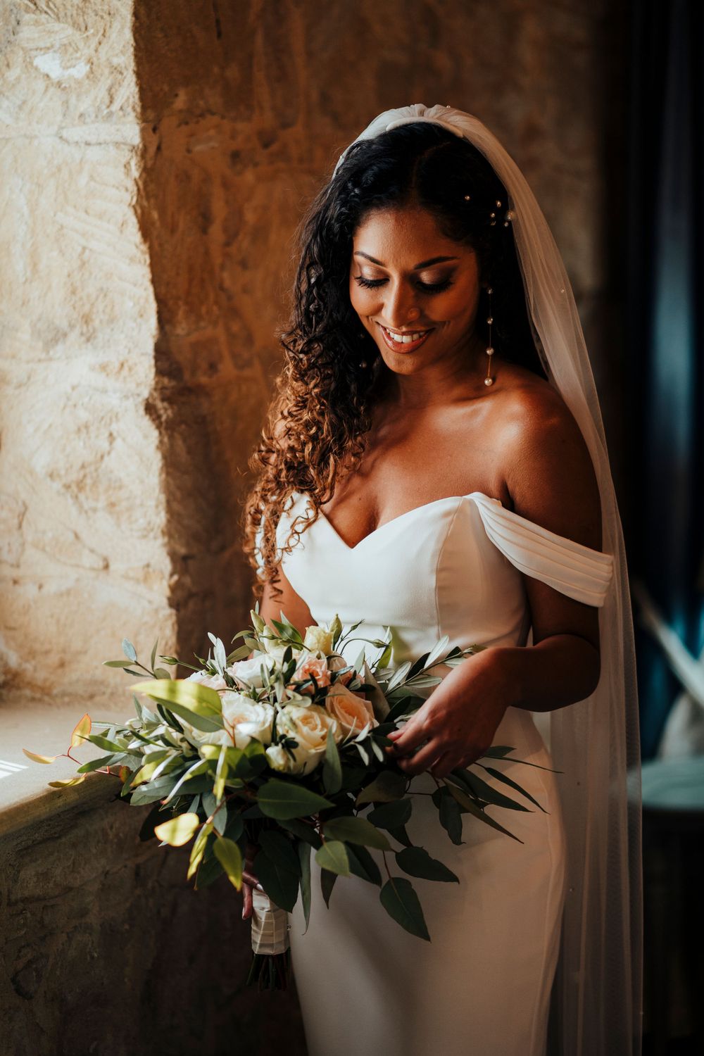 Bride with natural curls holds elegant white and green bouquet near rustic stone window.