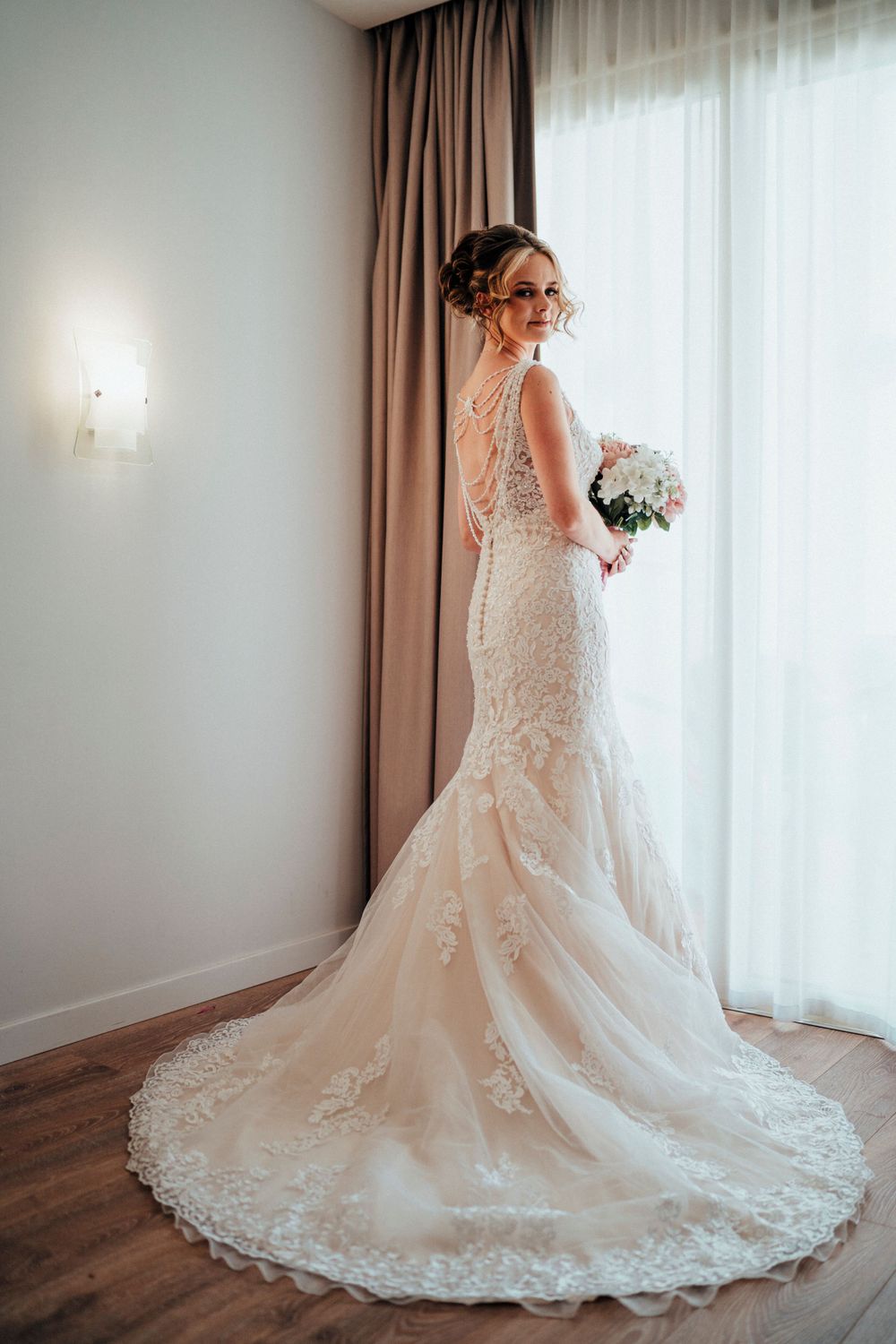 A bride in a fitted lace wedding dress with long train poses near a window in an elegant room.
