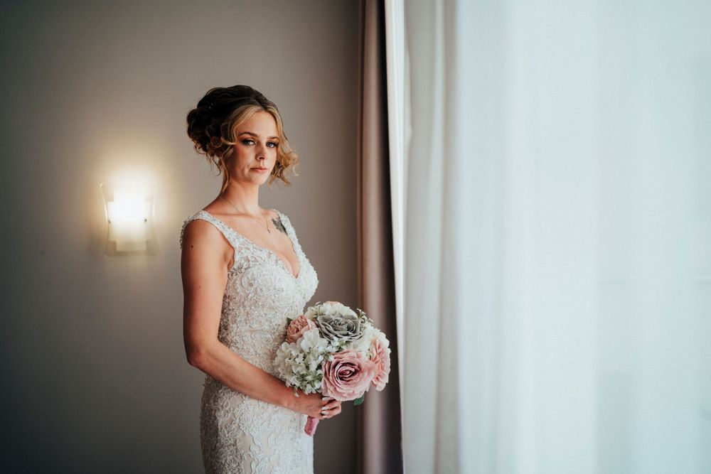 A bride holding a bouquet poses by a window in a fitted lace gown with natural lighting.