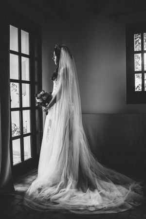 A dramatic black and white photo of a bride with long veil standing by vintage windows.