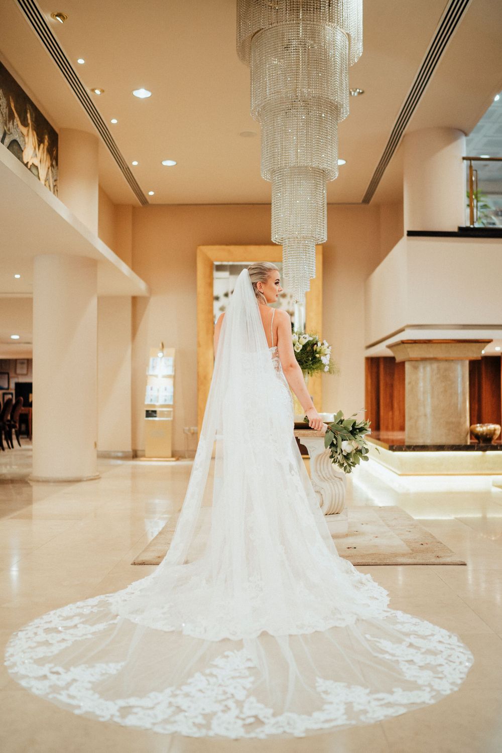 Dramatic cathedral length lace veil trailing on marble floor under crystal chandelier.