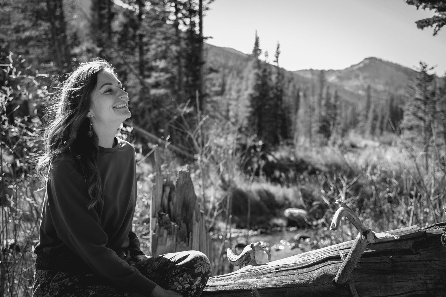 Person in dark clothing sits contemplatively on log in mountain wilderness.