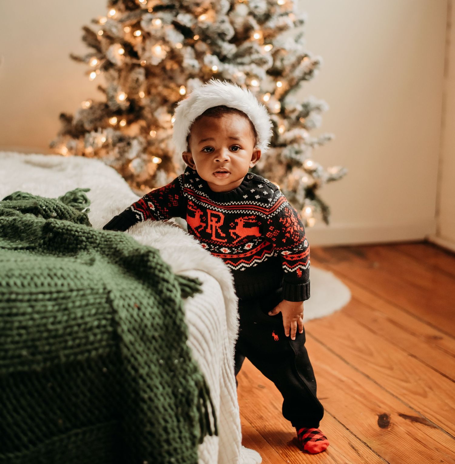 Adorable toddler in Christmas sweater and Santa hat stands by a decorated holiday tree.