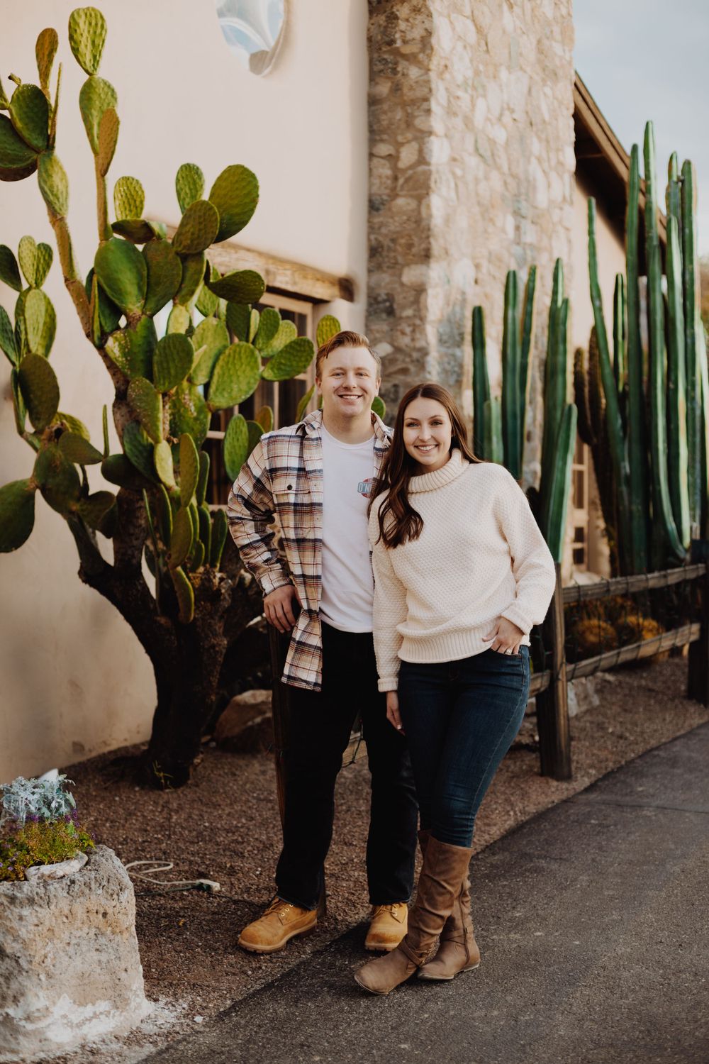 Couple stands together in front of desert landscaping with cactus plants and stone walls.