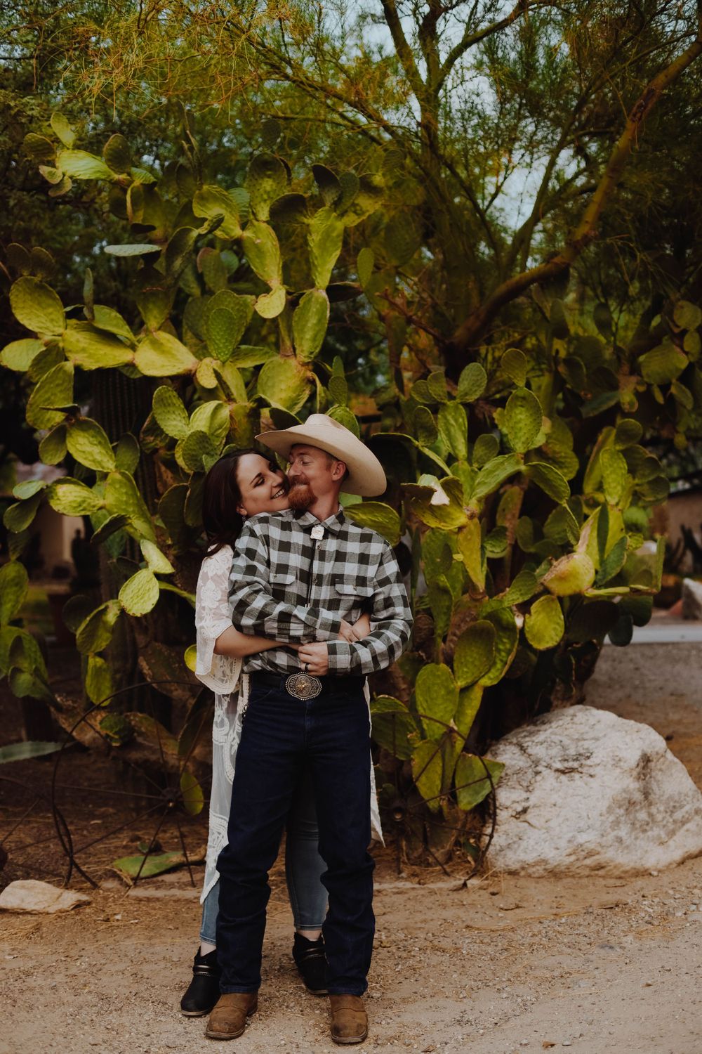 Couple embraces near large cactus plants in a desert garden setting.